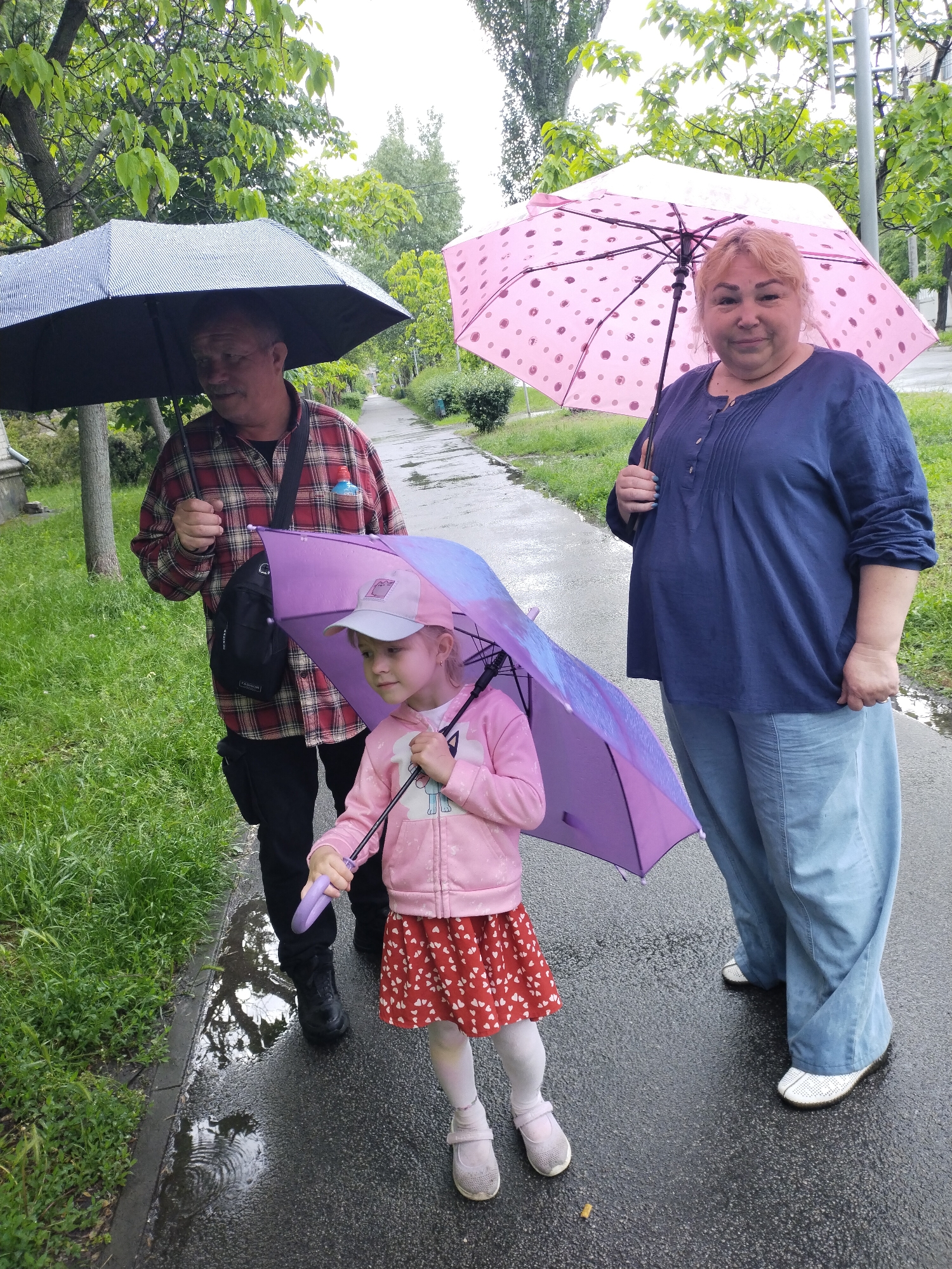 Three people stand on a wet sidewalk under umbrellas on a rainy day. A man with a dark umbrella and a red plaid shirt is on the left, a woman with a pink polka dot umbrella and a blue blouse and light blue pants is on the right, and a small girl with a purple umbrella, pink jacket, and red and white heart patterned skirt is in the center. The sidewalk is wet with puddles, and there is green grass and trees lining the sidewalk. The scene appears to be in a residential area. Based on visual context, this photo was taken in the city of Horishni Plavni, Ukraine.