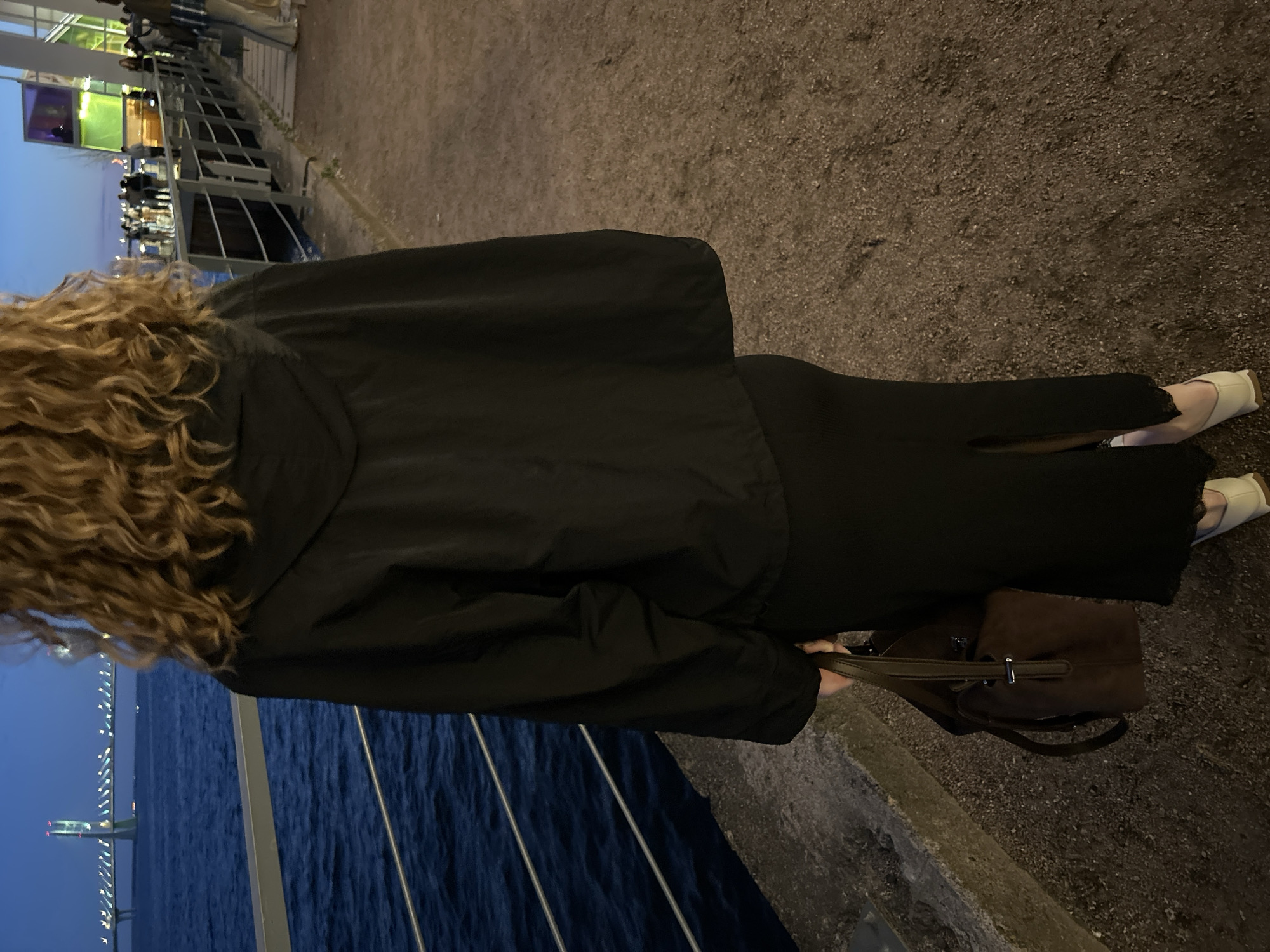 The image shows a person from behind, standing on a concrete path next to a body of water. The person is wearing a black hooded jacket and a long black skirt with a slit. They are carrying a brown handbag. In the background, there is a bridge with lights spanning across the water, and other people are visible on a walkway to the right. The sky is dark blue, indicating it is either dusk or night. The location is likely an embankment or promenade in Saint Petersburg, Russia, given the presence of a bridge and the architectural style of the building visible in the far background on the right, which resembles the New Holland Island cultural space.