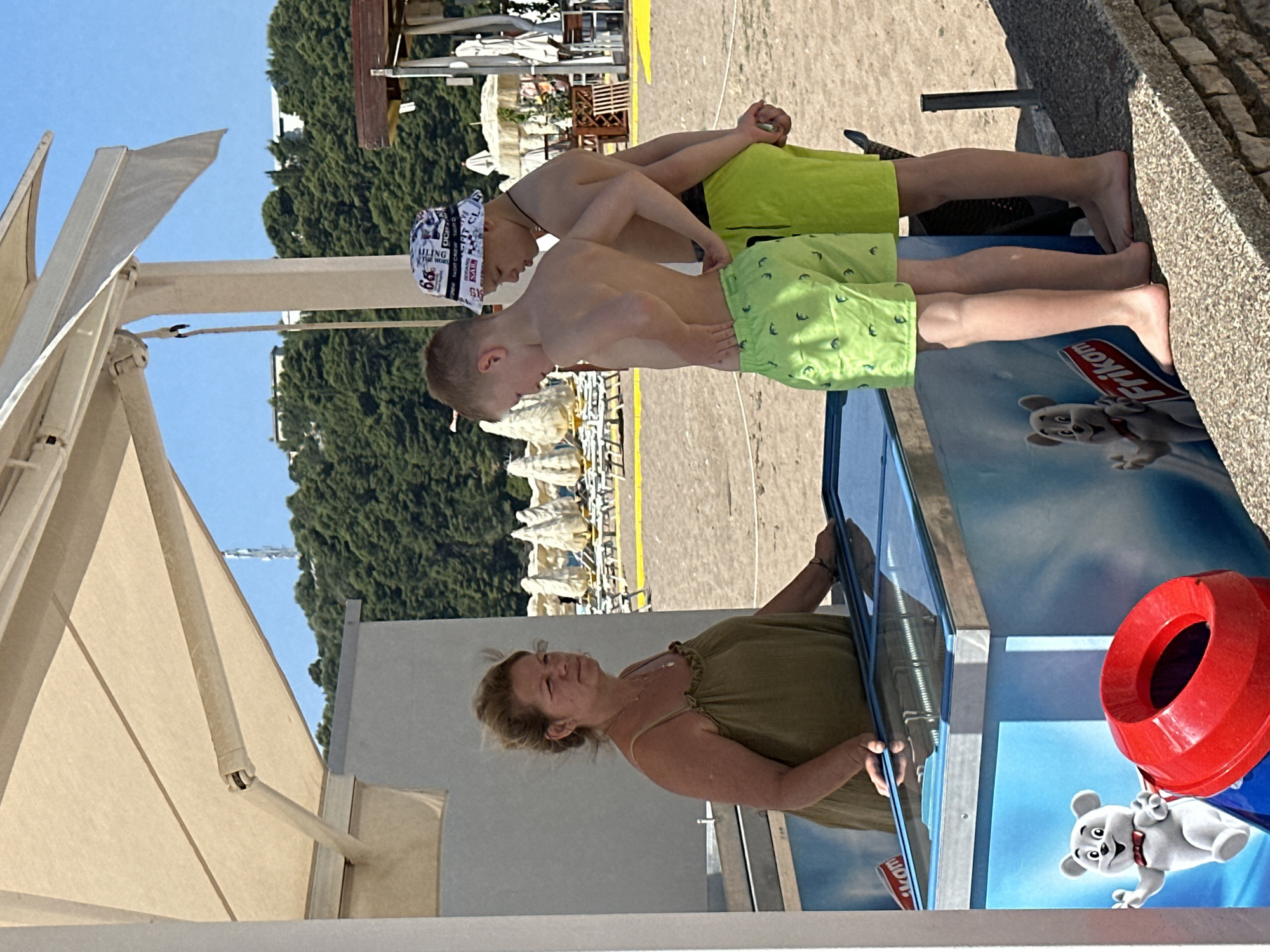 The image depicts a sunny day at what appears to be a beach or a beachside establishment in Boreti, Montenegro. In the foreground, a woman in a green dress is looking upwards and slightly to the right, with a subtle smile on her face. She is holding onto a blue reflective surface, possibly a window or a display case. Next to her, a red trash receptacle is visible.
Behind the woman, two young boys are standing. The boy in front is wearing light green shorts with a pattern of small dolphins. The boy behind him is wearing slightly darker green shorts and a white bandana with black text. Both boys are shirtless and appear to be interacting or posing.
The background suggests a beach environment with sand, lounge chairs, and umbrellas, indicating a leisure setting. Lush green trees are visible on a hill in the distance, under a clear blue sky. The lighting suggests it is daytime, likely midday due to the brightness and shadows. A partial view of a beige awning structure is also present in the left side of the image, providing shade. The overall atmosphere is relaxed and summery. Visible text includes "Frikom" on a blue surface near the bottom of the image, and fragmented text on the boy's bandana.
