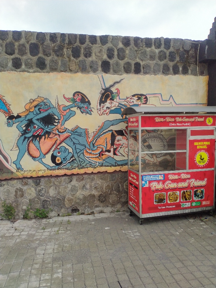 An outdoor scene in Karanganyar, Indonesia, features a vibrant mural painted on a textured, light-colored wall, which rests atop a rugged stone foundation. The mural, rendered in a traditional Indonesian style reminiscent of Wayang figures, depicts several blue-skinned characters engaged in what appears to be a struggle or dynamic interaction. One large, multi-armed figure dominates the left side, while other figures, some fallen or in motion, are spread across the mural. Beneath these figures, fragmented Indonesian text is visible, including words like "Remeng," "tidak," and "adalah."
In front of the mural, to the right, stands a bright red food cart on wheels. The cart has glass display cases, though they appear mostly empty. Prominently displayed on the cart is the branding "Rica - Rica Pak Gun and Friend," with a smaller subtitle "(Dulu Rica Fadhil)." Yellow labels on the side advertise "KULINER PEDAS BERLEVEL" (Spicy Culinary Level) and "KULINER INDONESIA BEDA RASA" (Indonesian Cuisine Different Taste), featuring a duck logo. Below, it notes "MITRA UMKM INDOMARET" (Indomaret MSME Partner) and shows images of various food items, including chicken/meat dishes and fried snacks. The bottom of the cart reads "Terima Pesanan" (Accept Orders).
The ground beneath the cart and wall is paved with gray rectangular blocks. The sky above is overcast, suggesting a cloudy day.