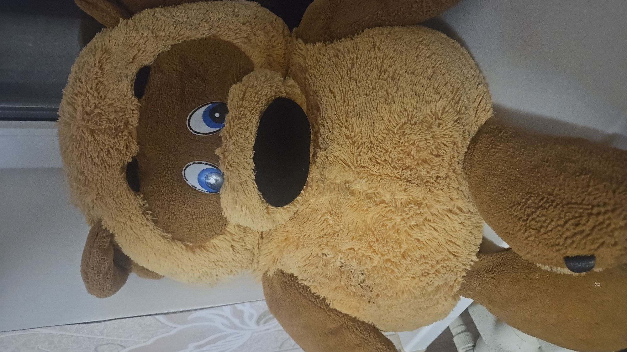A large, brown and tan stuffed animal, possibly a dog or bear, is captured in a close-up, rotated view. The plush toy features shaggy, light tan fur covering its main body and head, contrasting with darker brown fur on its limbs, ears, and snout. It has large, round blue eyes with distinct white reflections, giving them a glossy appearance. On its face, there's a prominent, oval-shaped black area, possibly representing an open mouth or a stylized dark muzzle. A smaller, distinct black nose is visible on the end of its snout.
The stuffed animal is positioned indoors, lying on a light-colored floor that appears to be tiled with a subtle, swirling pattern. A plain, light-colored wall or surface runs along the background, with a dark, indistinct object visible behind the animal's head. No specific activity is taking place; the animal appears to be resting or placed. There is no visible text in the image. The time of day and weather are not discernible from the indoor setting. The location context is Sevastopol, Ukraine.