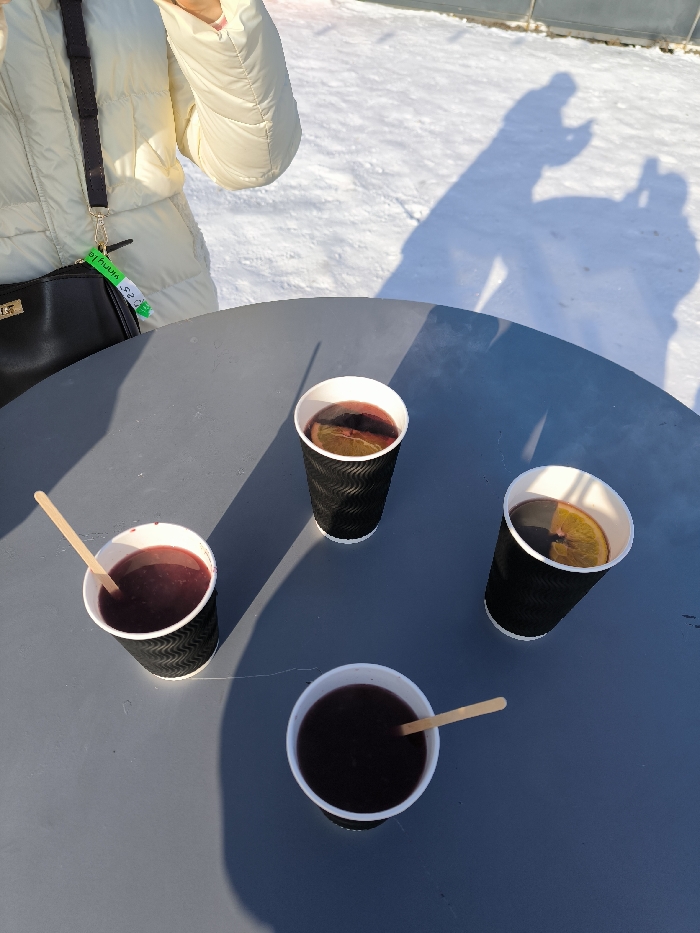 The scene is an outdoor winter environment in Prybuzke, Ukraine, under direct sunlight. A dark grey, circular table occupies the central and lower portions of the frame, holding four disposable cups. Two black, ridged cups, positioned on the right, contain a dark red beverage with an orange slice floating on the surface. Two other cups, located on the left (one black ridged, the other generic white with a black base), contain a similar dark red liquid, each featuring a small wooden stirring stick.
On the upper left, a person is partially visible, wearing a cream-colored puffy winter jacket. This individual is carrying a black shoulder bag, from which a green and white rectangular keychain or tag, partially labeled "AULIN" and "SES", is suspended. The background consists of a snow-covered ground, which appears textured and uneven. Long, distinct shadows of at least two individuals are cast onto the snow in the upper right quadrant of the image; one shadow suggests a person holding a device consistent with a camera or mobile phone.