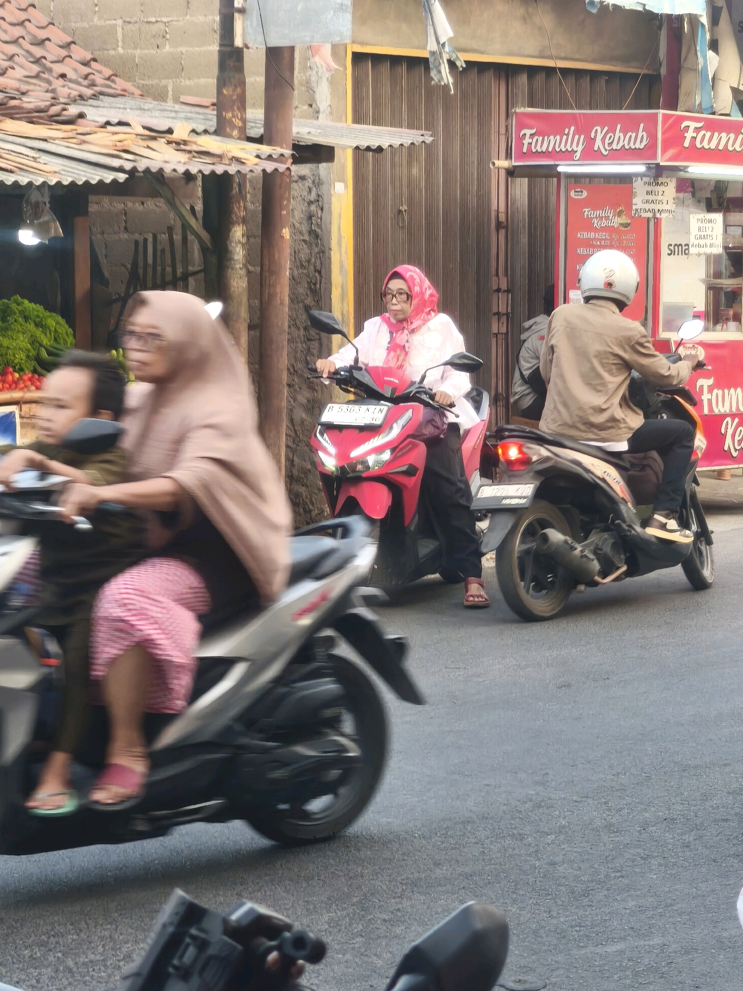 In Satriamekar, Indonesia, on a paved road in front of a building with a corrugated metal roof and brown sliding doors, three individuals are depicted on two motorcycles. The foreground shows a motorcycle with two passengers, a woman wearing a brown hijab and a pink and white patterned skirt, and a child in a dark shirt. The woman is driving, and the child is seated behind her. To the right, a second motorcycle is occupied by a person in a light brown jacket and a white helmet, also driving. In the background, a red and white sign reads "Family Kebab." The scene appears to be during daylight hours, with visible street elements and market stalls.
