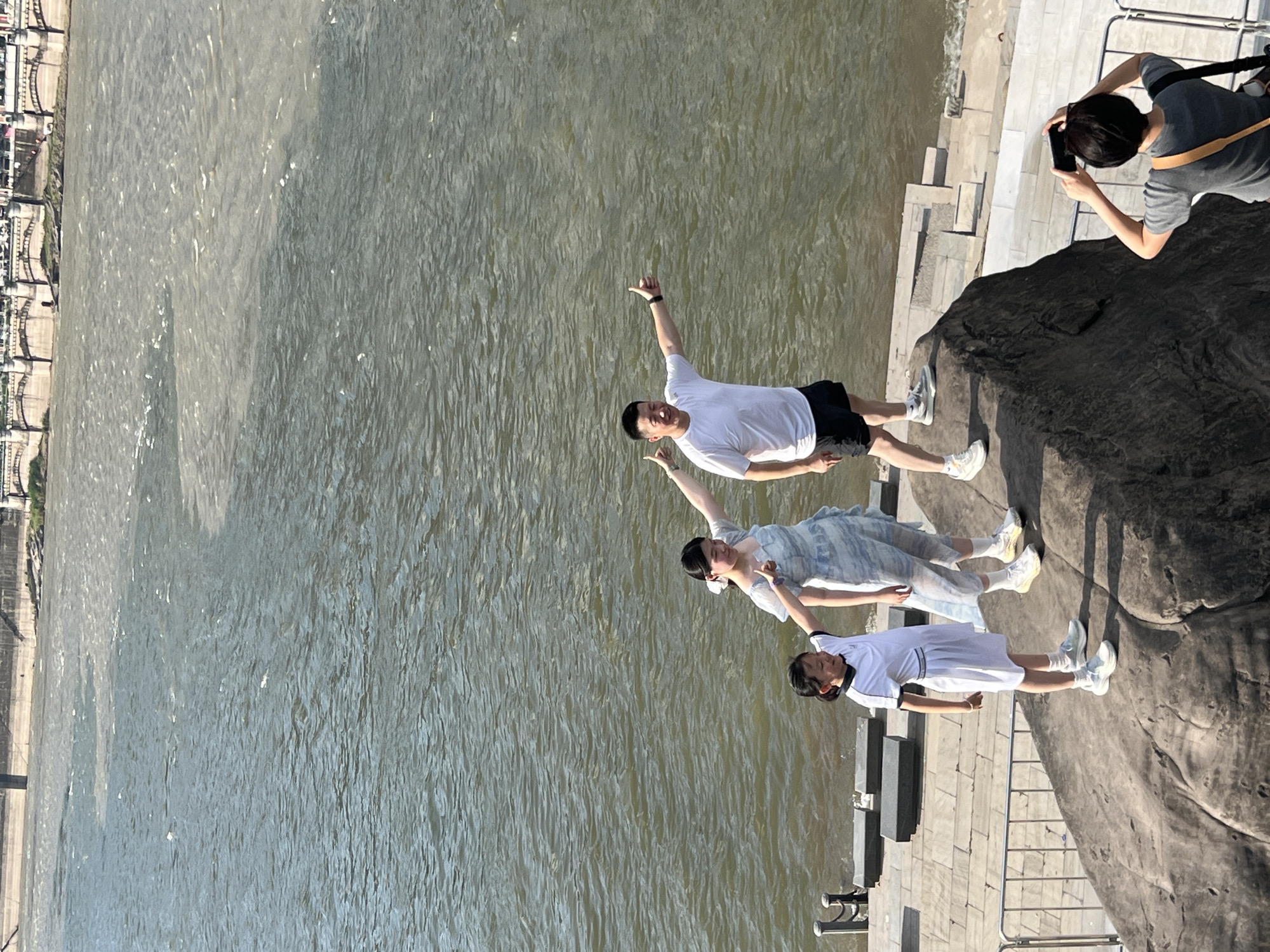 An outdoor, high-angle, medium shot captures three people posing on a large rock formation near a river in the Yuzhong District of China. A young girl in a white dress stands on the left, looking at the camera and smiling. Beside her, a woman in a light blue dress also looks towards the camera, with her left arm extended. A man in a white t-shirt and black shorts stands on the right, giving a thumbs-up to the camera. In the foreground, a person with dark hair is partially visible, taking a photo of the three with a black smartphone. The river behind them shows a distinct line where two different colored bodies of water meet. A bridge and a stone embankment are visible in the background, along with a city skyline. The overall scene suggests a tourist activity.