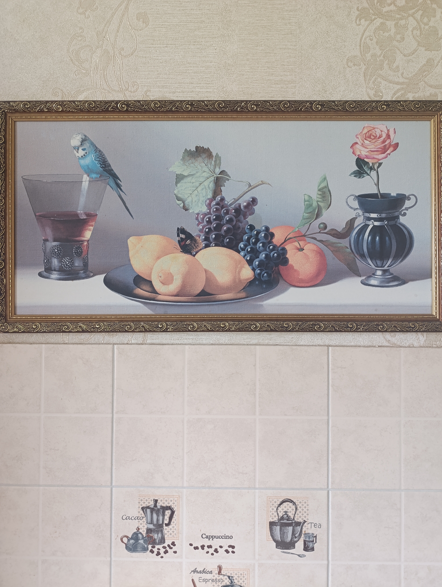 The image shows a still life painting of fruits, a bird, and a rose in a vase. The painting is framed in ornate gold-colored wood and hangs on a beige textured wallpaper. Below the painting are beige square tiles. On the tiles, there are several small decorative images related to coffee and tea, including a moka pot, a teapot, and text labels like "Cacao," "Cappuccino," "Tea," "Arabica," and "Espresso." The scene appears to be an interior domestic setting. The location is in the city of Bila Tserkva, Ukraine. There are no people present in the image.