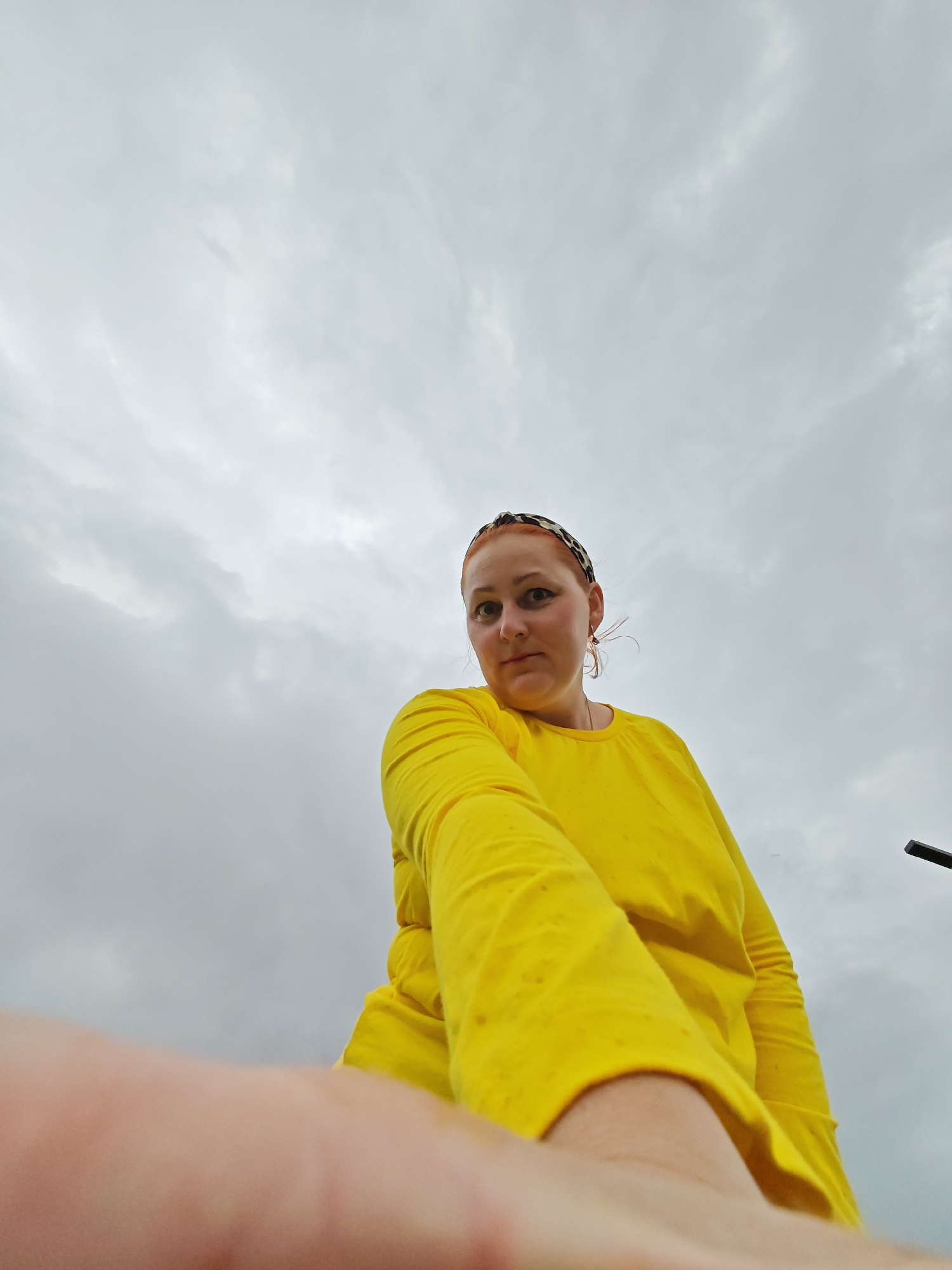 A woman with red hair is wearing a yellow long-sleeved shirt and a leopard-print headband. She is looking down at the camera, and her right arm is outstretched towards the camera. The background is a cloudy sky. The location is Марковский, Russia.