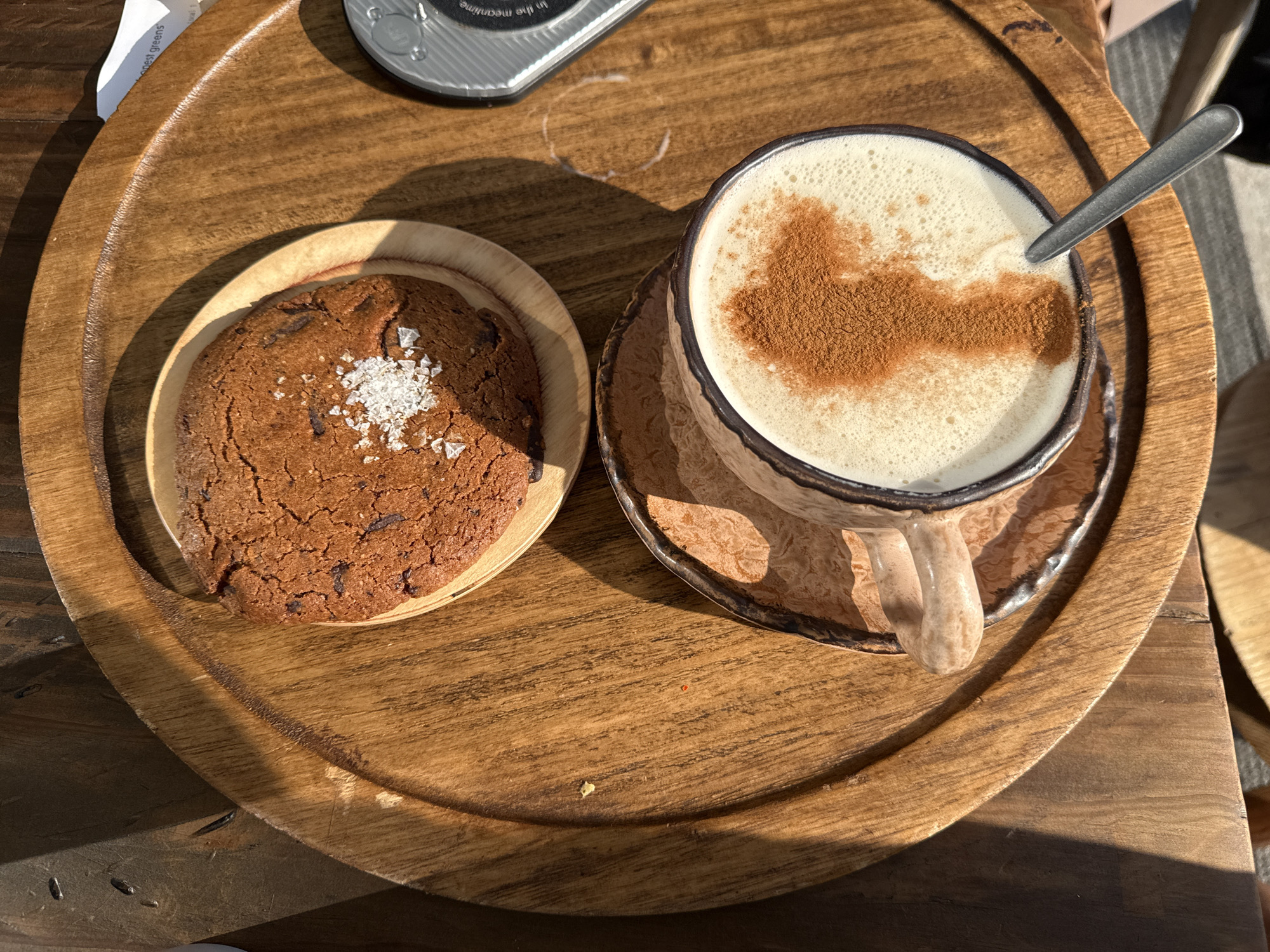 A rustic wooden tray holds a coffee mug with a marbled design and a frothy beverage topped with cinnamon, alongside a chocolate chip cookie sprinkled with sea salt. A silver spoon rests in the mug, and a white coaster with text is partially visible. The scene is bathed in natural light, casting shadows across the wooden surface. The location is Unknown, Unknown.