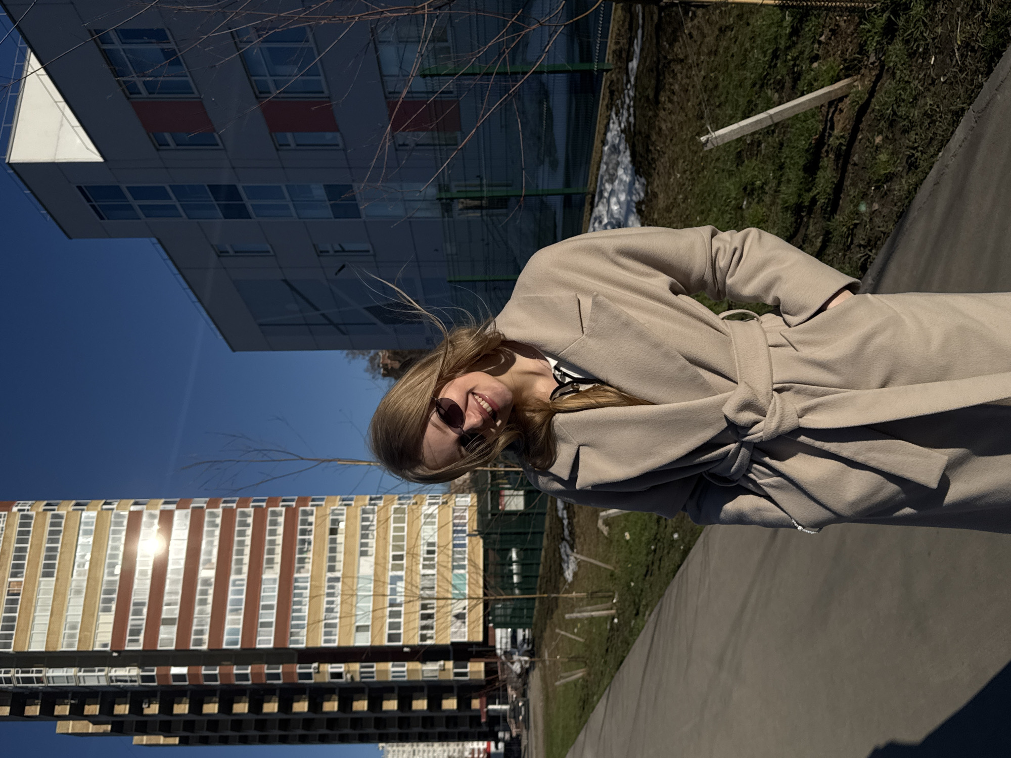 A young woman is standing on a sidewalk in Perm, Russia, smiling at the camera. She is wearing a light beige coat tied at the waist and sunglasses. Behind her is a grass area with some snow patches, and a large apartment building with alternating brown and beige vertical stripes on the facade. Further back, a smaller modern building with a white facade and red accents is visible. The sky is clear and blue.