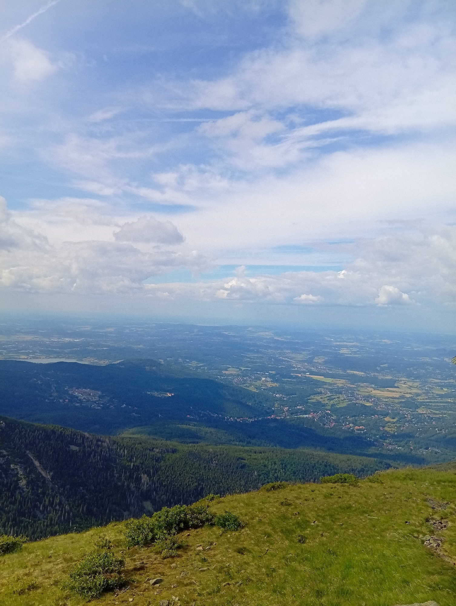 The image is a panoramic view from a mountain summit overlooking a valley and distant landscape. The foreground features a grassy slope with scattered rocks and low-lying green bushes. In the mid-ground, a dense, dark green forest covers the mountainside, with some lighter green patches and visible tree tops. Further into the valley, a scattering of buildings and settlements are visible, interspersed with fields of varying colors, suggesting agricultural activity. The background reveals a hazy, expansive landscape stretching to the horizon under a partly cloudy blue sky. The scene depicts a natural environment with signs of human habitation in the valley below. This view is likely from a location in or near Malá Úpa, Czechia, offering a vista of the surrounding countryside.
