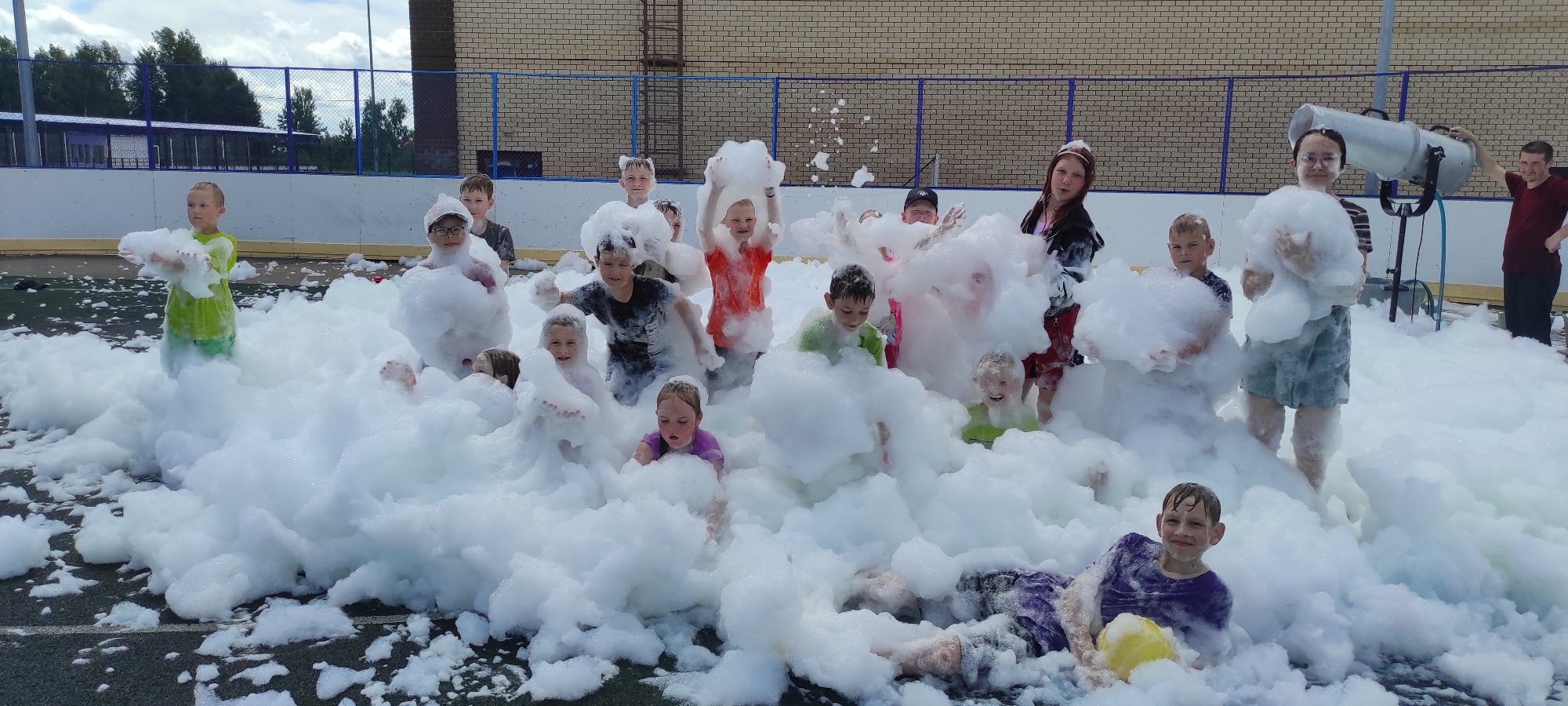 A group of children are playing in a large pile of white foam, likely from a foam machine, in an outdoor setting that appears to be a sports court or playground in Noviy, Russia. The children are of various ages, mostly dressed in summer clothing, and are actively engaged with the foam, some holding it, others submerged in it, and some looking towards the camera. In the background, there is a building with brick walls and a chain-link fence. A foam machine is visible on the right side of the image, operated by an adult. The sky is cloudy.