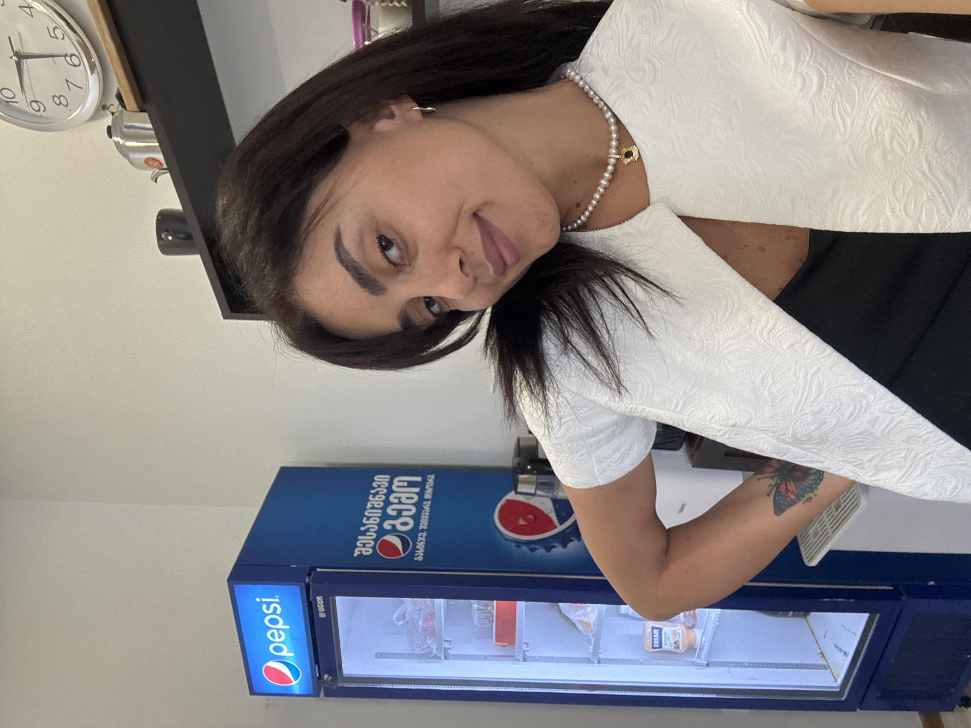 A woman with dark hair and brown eyes poses in front of a Pepsi refrigerator. She is wearing a white textured jacket over a black top and a pearl necklace with a dark pendant. On her left arm, she has a butterfly tattoo. The refrigerator has the Pepsi logo on top and text in Georgian. In the background, a shelf holds a clock, a kettle, and a cup. The scene appears to be indoors, possibly in an office or commercial space in Tbilisi, Georgia.