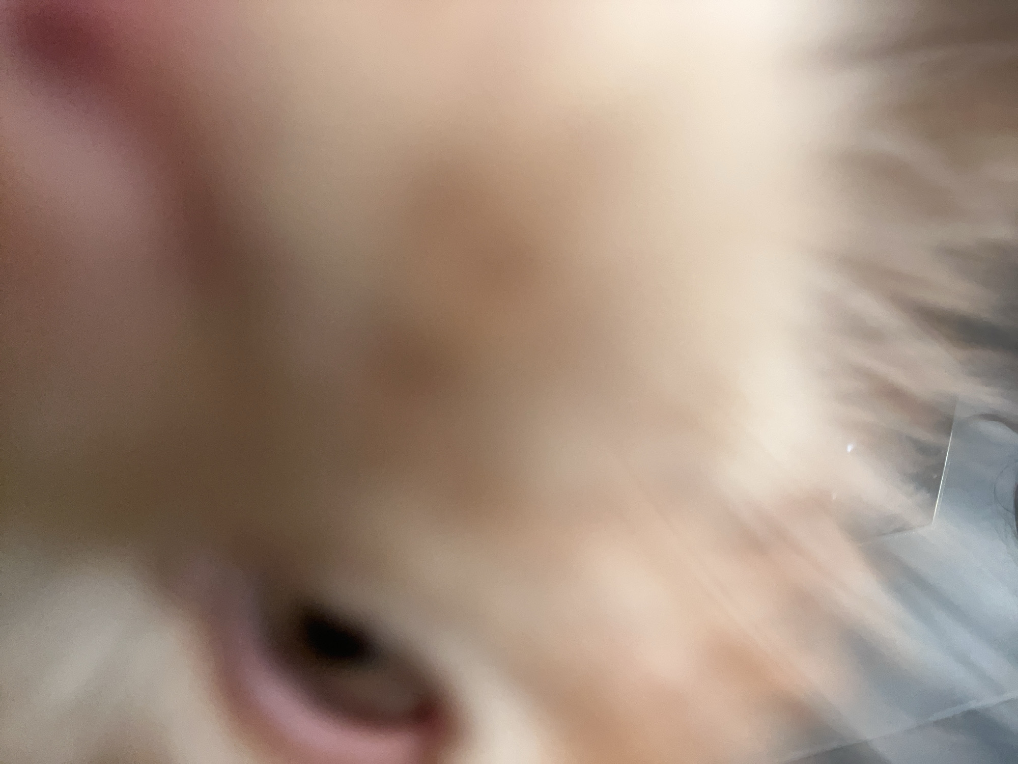 A close-up, out-of-focus, low-angle shot captures a blurry, light-colored subject in the foreground. The subject appears to be a pet, possibly a cat or dog, with its fur exhibiting shades of beige and white. The image is dominated by soft, diffused light, creating a dreamy and indistinct atmosphere. A hint of a dark eye and a pink ear are visible on the right side of the frame, suggesting the face of the animal. The background consists of blurry, cool-toned streaks of light and shadow, possibly indicating a window or a reflective surface. The overall impression is one of an intimate and personal moment, focusing on the texture and form of the animal's features.
