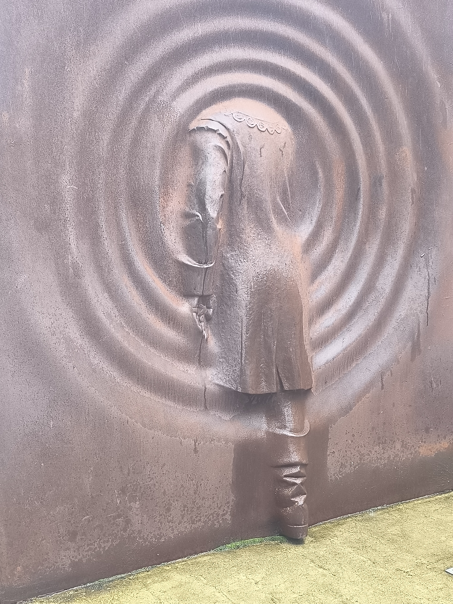 The image shows a textured, reddish-brown metal sculpture of a figure with its head bowed and turned away from the viewer. The sculpture is set against a backdrop of concentric, ripple-like patterns also etched into the metal. The figure appears to be wearing clothing that drapes and folds, with a distinct section resembling a boot or gaiter on its visible leg. The sculpture is positioned outdoors, with a yellow-ish, textured ground surface visible at the bottom, bordered by a thin strip of green. This sculpture is located in Kuldīga, Latvia, at the site of the Venta Waterfall.