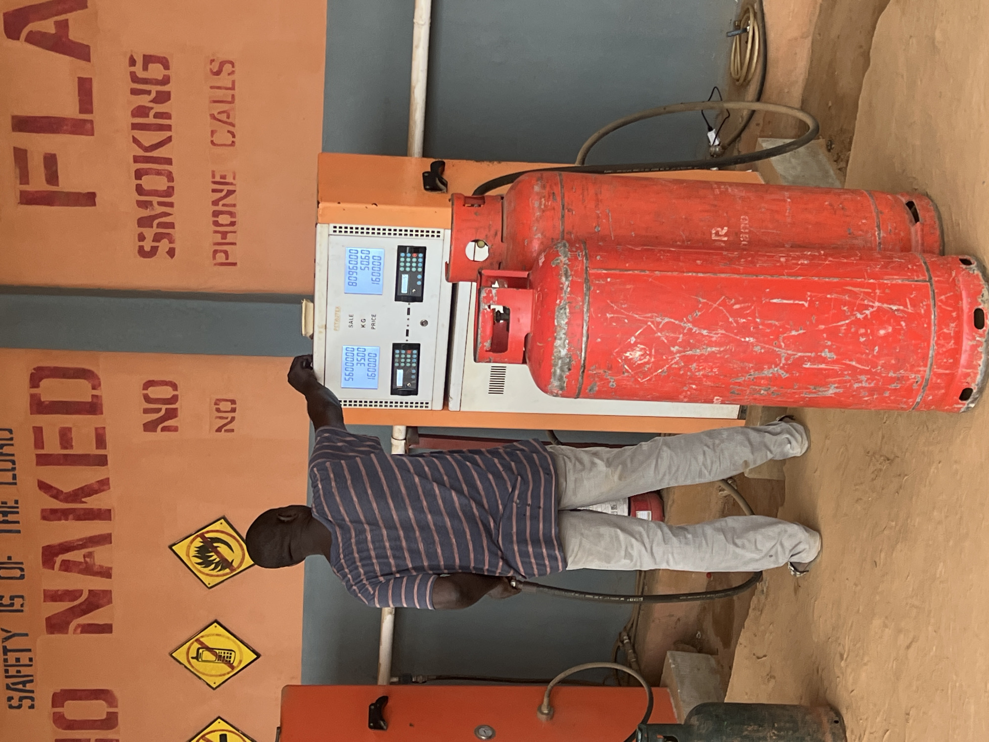 The image depicts a fuel station with gas cylinders in Nkpor, Nigeria. In the foreground, a man wearing a striped t-shirt and grey trousers is standing at a fuel dispenser, holding a nozzle. The dispenser has digital displays showing numbers that likely represent fuel sales and prices. Behind the man, two large red gas cylinders are visible, along with a green one on the left. The wall behind the dispenser is painted orange with red stencil text that reads "SAFETY IS OF THE LORD", "NO NAKED", "NO SMOKING", and "PHONE CALLS". There are also two yellow hazard signs with red symbols on the wall. The ground is covered in reddish-brown dirt, suggesting an outdoor setting. The overall scene suggests a functional, yet somewhat weathered, fuel station.