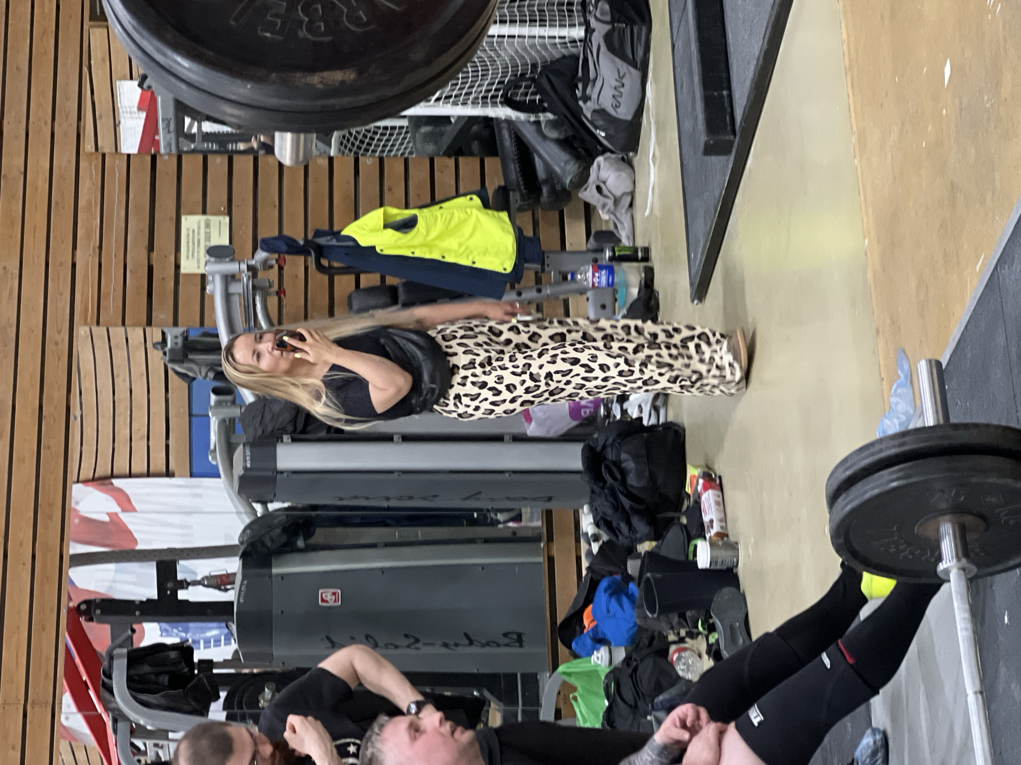 A gym interior is shown. A woman with long blonde hair is talking on her cell phone, dressed in a black top and leopard print pants. A man is lying on his back, seemingly preparing to do a leg press exercise with weights loaded on the machine. Other gym equipment is visible in the background. The location appears to be an indoor gym. There is no direct evidence pointing specifically to Saint Petersburg, Russia.