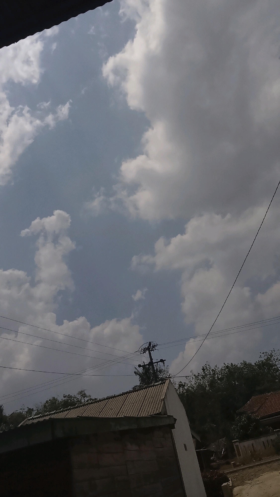 The image depicts a cloudy sky above a rural scene in Bojong Barat, Indonesia. In the foreground, a portion of a dark, possibly wooden structure is visible, with a corrugated metal roof extending to the right. Beyond this, a white building with a sloped roof is partially seen. Several utility poles and electrical wires crisscross the mid-ground, with trees and foliage lining the horizon. In the far right, a roof with red tiles suggests another dwelling. The sky is a mix of blue patches and white and gray clouds, indicating partly cloudy weather. The overall impression is of a slightly overcast day in a residential area with visible infrastructure.