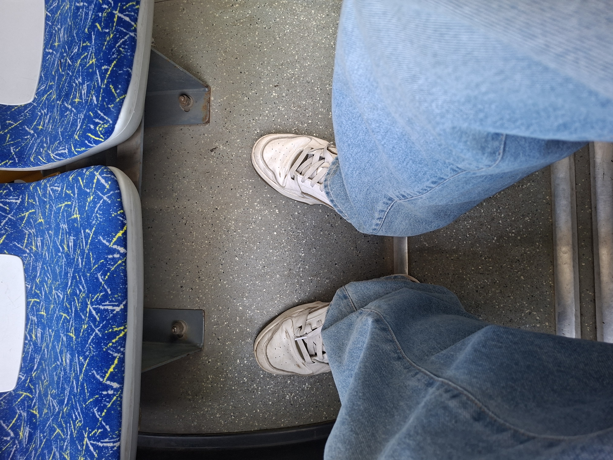 The image shows a first-person perspective of someone sitting on a public transport seat, likely a bus or train, with their legs and feet visible. The seats are upholstered in blue fabric with a pattern of white and yellow squiggles. The floor is a speckled grey material. The person is wearing light blue denim jeans and white, worn sneakers. The background consists of the seats in front and the floor. There is no indication of the location being Kazan, Russia.