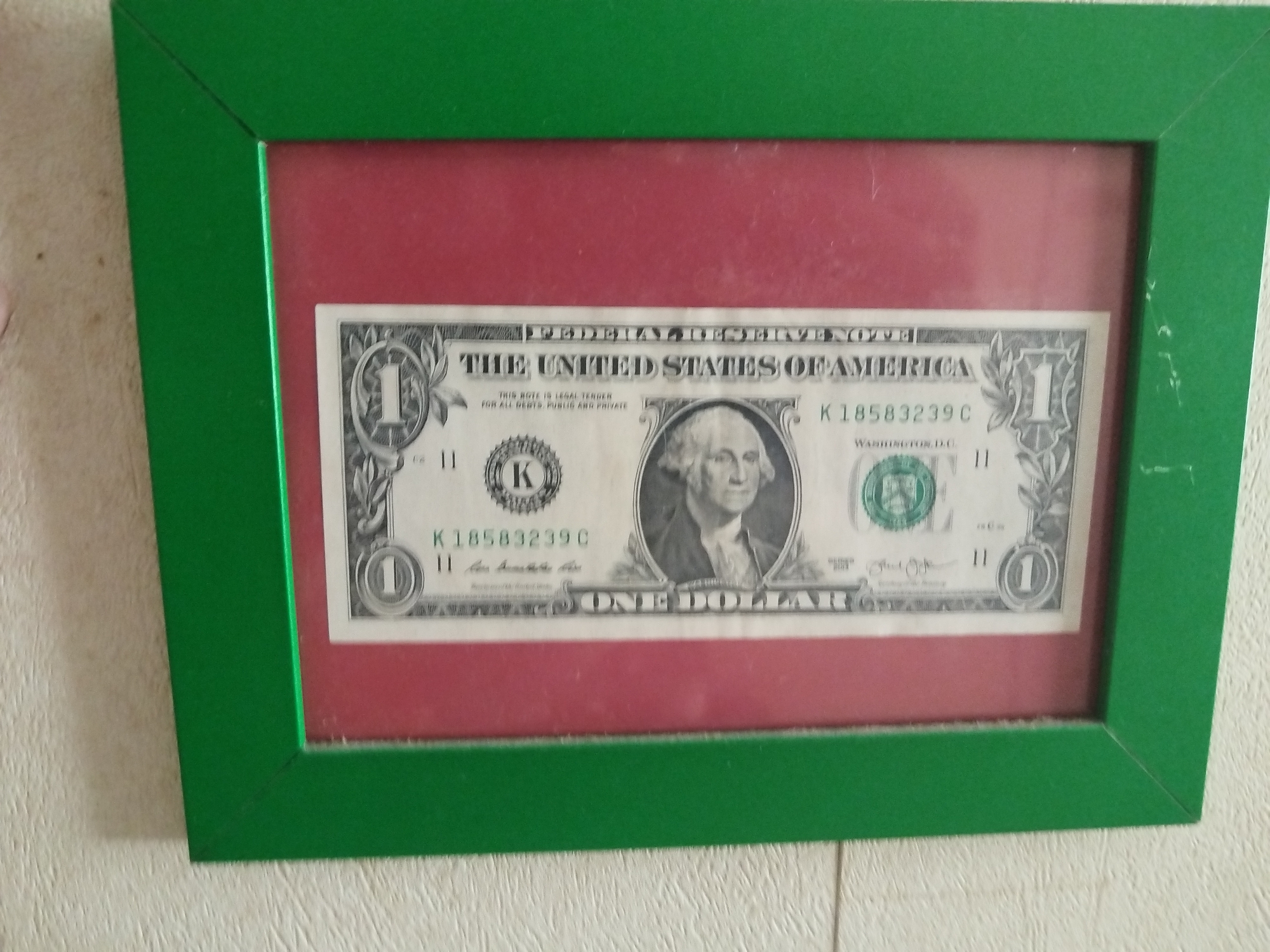 A framed one-dollar bill is hung on a textured white wall. The frame is green, and the backing inside the frame is red. The dollar bill is centered and displays the image of George Washington. The serial number on the bill is K18583239C. There is no information to suggest the picture was taken in the city of Baranovka, Russia.