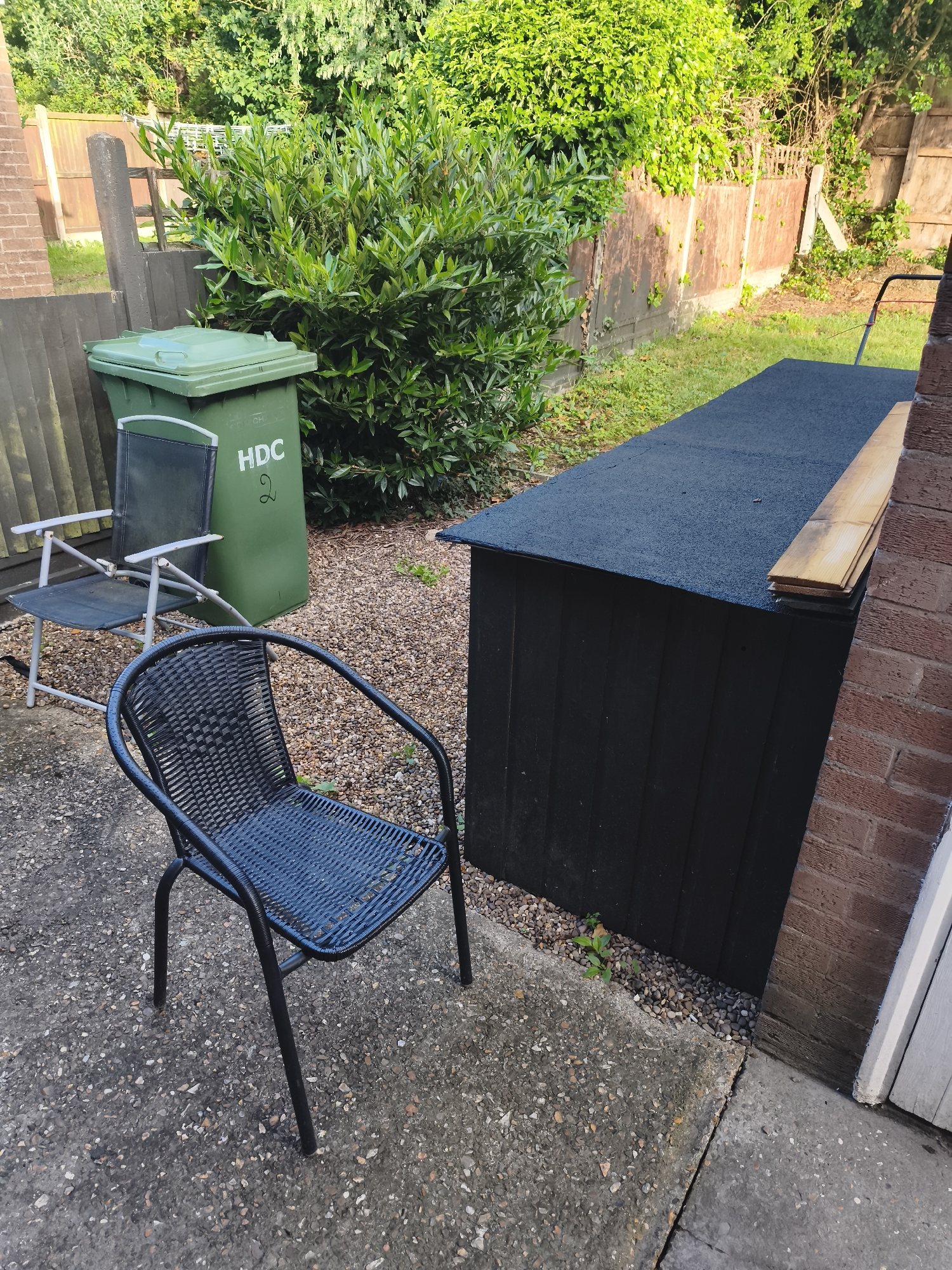 The image captures an outdoor domestic scene, likely a patio or garden area in Huntingdonshire, United Kingdom. In the foreground, a dark, woven-style garden chair with metal legs is placed on a concrete surface. Behind it, to the left, is a lighter-framed folding chair with dark fabric, situated on the same concrete area. Adjacent to the folding chair stands a green wheelie bin, prominently displaying the white text "HDC 2" on its side.
To the right of the chairs and bin, a long, low-profile storage unit or shed, painted black, occupies a gravel-covered patch. Its flat roof is also black and textured. A small stack of light-colored wooden planks rests on top of this unit, positioned against a brick wall that forms the far-right boundary of the image. The gravel area extends behind the bin and storage unit, leading towards a lush green bush with dense foliage that acts as a natural backdrop.
Behind the bush and to the left, a wooden fence runs along the perimeter, partially obscured by the greenery. Another section of wooden fencing is visible behind the bin. The scene is illuminated by bright natural daylight, casting some shadows and highlighting the vibrant green of the plants. A handle, possibly from a lawnmower, is just visible in the upper right background, near the fence line. The overall impression is a functional and lived-in outdoor space.