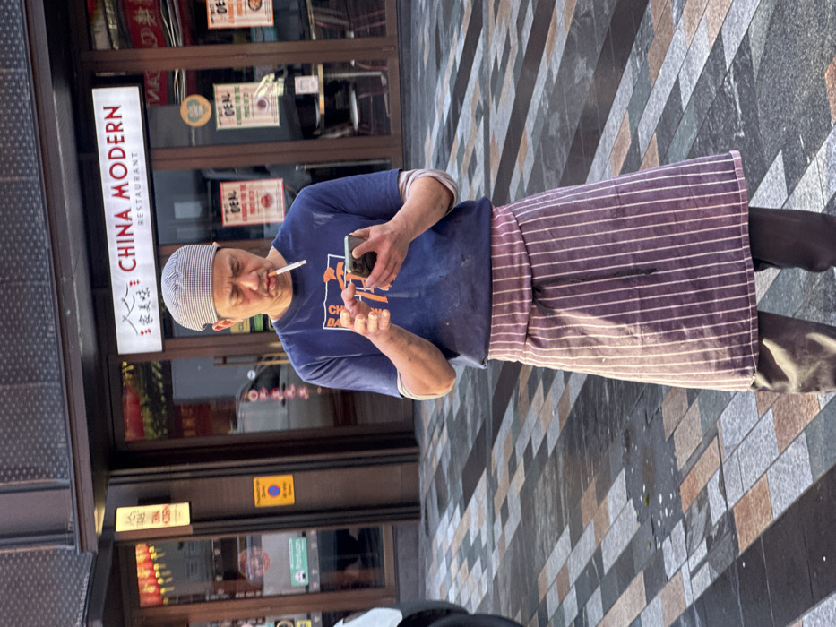 An East Asian male, appearing middle-aged, stands outdoors on a paved sidewalk, identified as Wardour Street, in London's Chinatown. He wears a checkered skull cap, a soiled dark blue t-shirt with an orange graphic, a vertically striped maroon apron, and dark trousers. A lit cigarette is positioned between his lips. He holds a black smartphone horizontally with both hands, looking down and interacting with its screen using his right thumb. Behind him is the storefront of "CHINA MODERN RESTAURANT," featuring prominent English and Chinese signage, glass doors, and visible interior elements including tables, chairs, and "DEAL" promotional posters. Red lanterns are visible inside the left window. The ground is paved with rectangular tiles in varying shades of grey and brown. A partial white vehicle is visible on the far left.