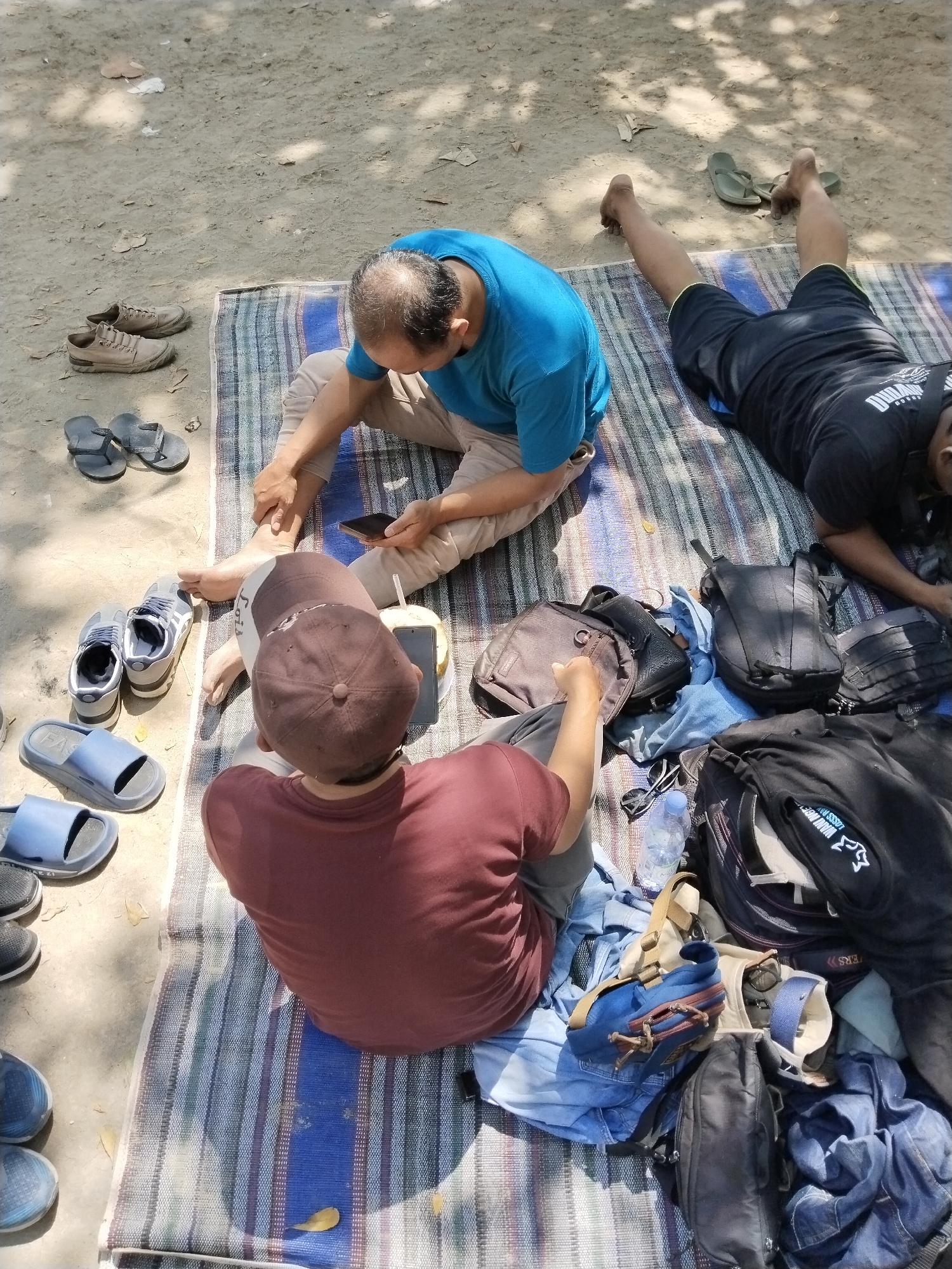 In Prigi, Indonesia, two individuals are seated on a striped mat outdoors. The individual on the left, wearing a blue t-shirt and tan pants, is looking down at a smartphone. The other individual, wearing a maroon shirt and brown cap, is facing away from the camera, looking down as well. Scattered around them are various items including shoes, sandals, backpacks, clothing, a water bottle, and a coconut. The ground is sandy with some dried leaves and debris. The scene appears to be under dappled shade from overhead foliage.