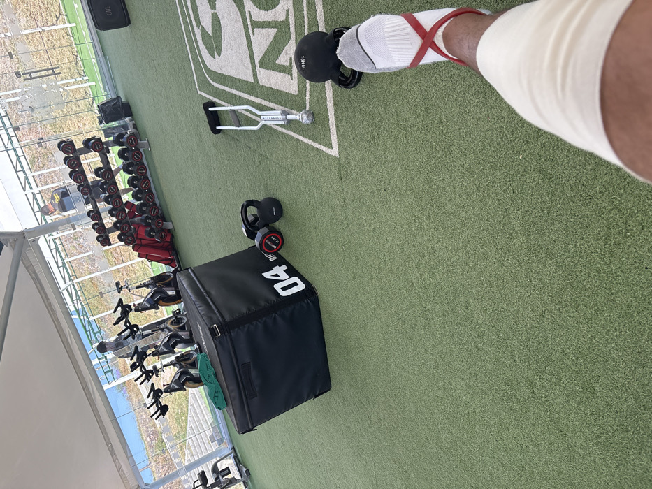 The image captures a first-person perspective, looking down at a person's leg and foot within what appears to be an outdoor or semi-outdoor gym facility.
In the immediate foreground, to the right, is a person's lower right leg and foot. The foot is clad in a white sock with grey stripes and accents. A red resistance band is wrapped around the ankle, crossing over the top of the foot. The lower leg, above the ankle, is encased in a light-colored, possibly beige or off-white, bandage or compression wrap. The person's skin tone is dark. The foot is positioned next to a black kettlebell, which prominently displays "16KG" in white lettering, indicating its weight.
The floor of the facility is covered in green artificial turf, resembling a sports field, with white markings forming part of a larger logo or text, where "S N" is visible.
Scattered across the artificial turf are various pieces of gym equipment. Near the person's foot, there is a silver or chrome piece of equipment, possibly a type of handle or part of a functional training apparatus, with a ribbed grip section. To the left of this, a black plyometric box (or jump box) is positioned, with the number "04" clearly marked in white on its side. Adjacent to the plyometric box are two smaller black weights, one of which shows "2.5KG" and "BRV" written on it.
In the middle ground and background, more gym equipment is arranged. Along the left side of the image, a rack of dumbbells with red handles and black weights is visible, ranging in size. Further behind the dumbbell rack, several stationary bikes or spin bikes are lined up.
The setting appears to be an open-air or partially enclosed gym, possibly under a large white tent or canopy structure, with support beams visible on the far left. Through glass panels or an open wall in the background, a view of an outdoor landscape can be seen, including what looks like a body of water (possibly a lake or ocean) and some buildings in the distance. The lighting suggests it is daytime, with bright natural light illuminating the area. In the upper left corner, a black bag or speaker with "JBL" written on it is partially visible.
The scene conveys an atmosphere of training or rehabilitation, given the specialized equipment and the bandage on the leg.