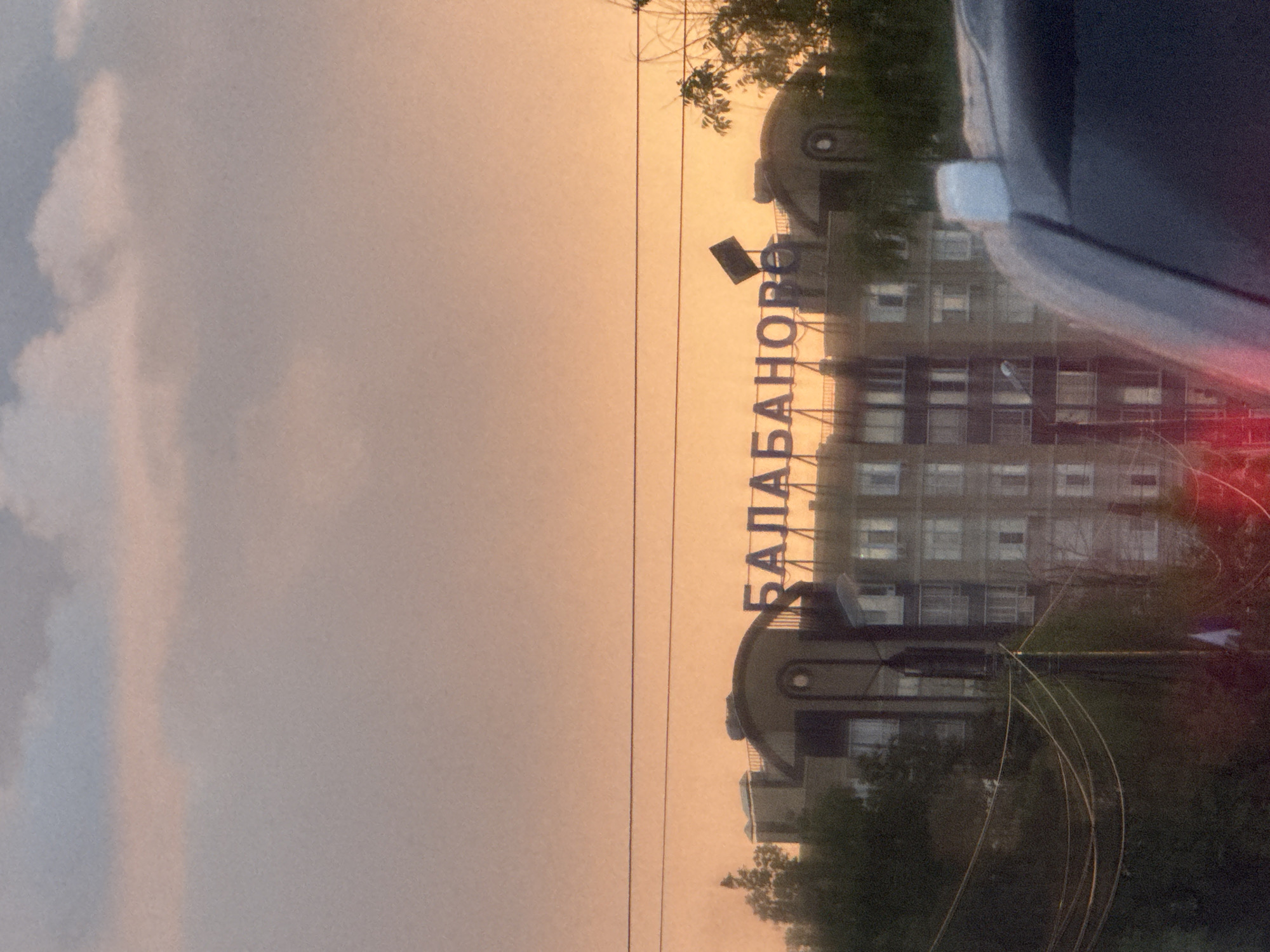 The image shows the entrance sign of the city of Balabanovo, Russia. The sign, spelling "БАЛАБАНОВО" in Cyrillic, is mounted on a building. The sky is a mix of clouds and an orange hue, suggesting either sunrise or sunset. In the foreground, the edge of a vehicle is visible. Utility lines also traverse the image. The overall scene appears to be an urban environment.
