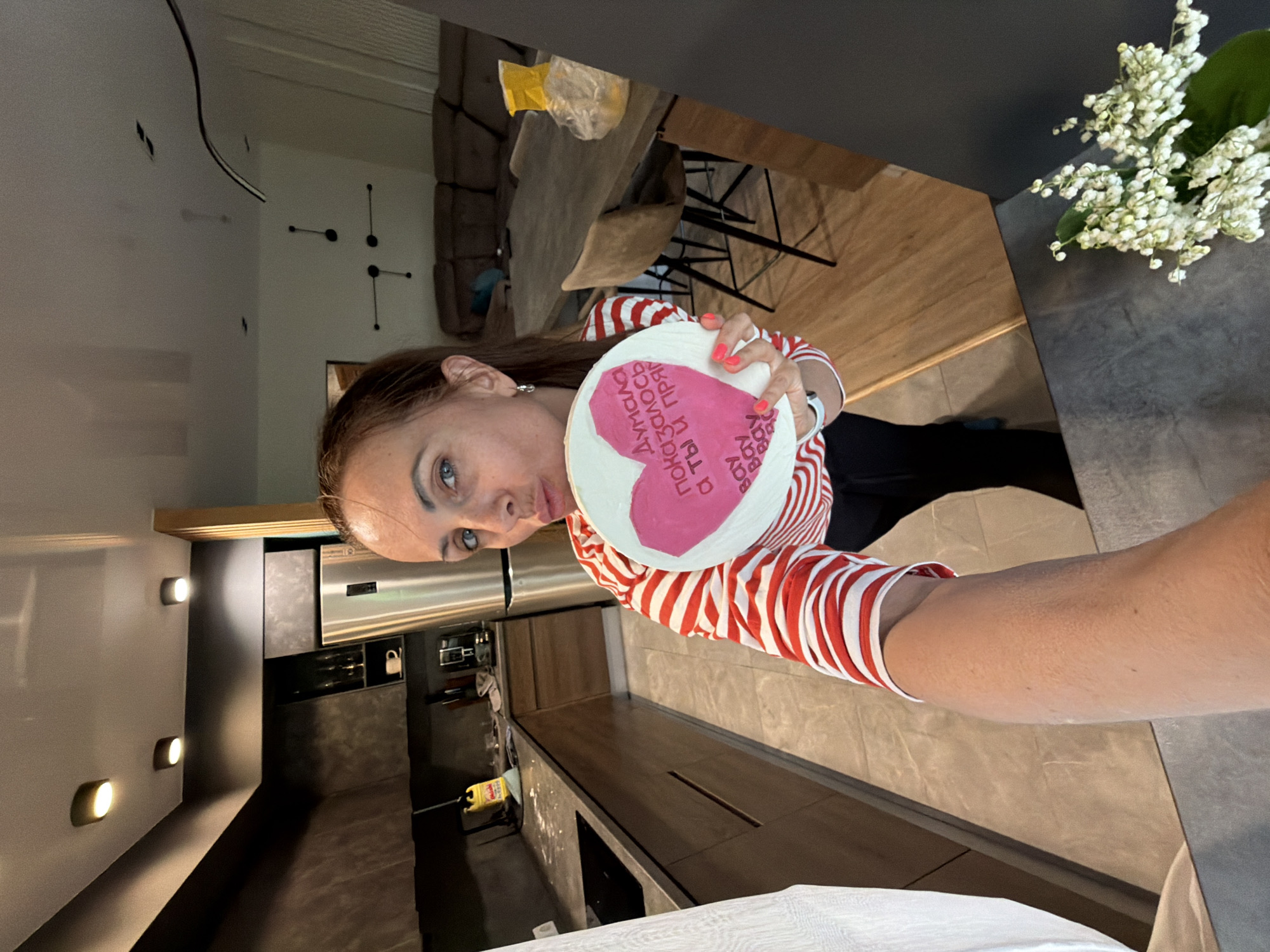 The image, taken in Birobidzhan, Russia, captures a woman taking a selfie in a modern, open-plan indoor setting that combines a kitchen, dining, and living area. The woman, with brown hair, blue eyes, and light skin, wears a red and white striped long-sleeved shirt and has bright pink nail polish. She looks directly at the camera with a somewhat serious or pouting expression.
She holds a round, white disc or plate adorned with a large pink heart. Within the heart, Cyrillic text is written. Partially visible, the text includes "Я ДУМАЛА" (I thought), "ПОКАЗАЛОСЬ" (it seemed/turned out), and "А ТЫ И ПРАВ" (and you are right). Below the heart, the word "ВСЁ" (everything/all) appears to be repeated. This text likely conveys a personal or humorous message.
The background shows a contemporary interior. To the right, a dark countertop holds a bouquet of white flowers, resembling lilies of the valley. Further back, a wooden dining table with chairs and a brown sofa are visible. A minimalist black geometric wall decoration is mounted on a light-colored wall. The kitchen area features dark countertops, modern cabinetry, stainless steel appliances, and wall-mounted lights, contributing to a clean and stylish aesthetic. The flooring transitions between wood and tile. The lighting suggests it is daytime.