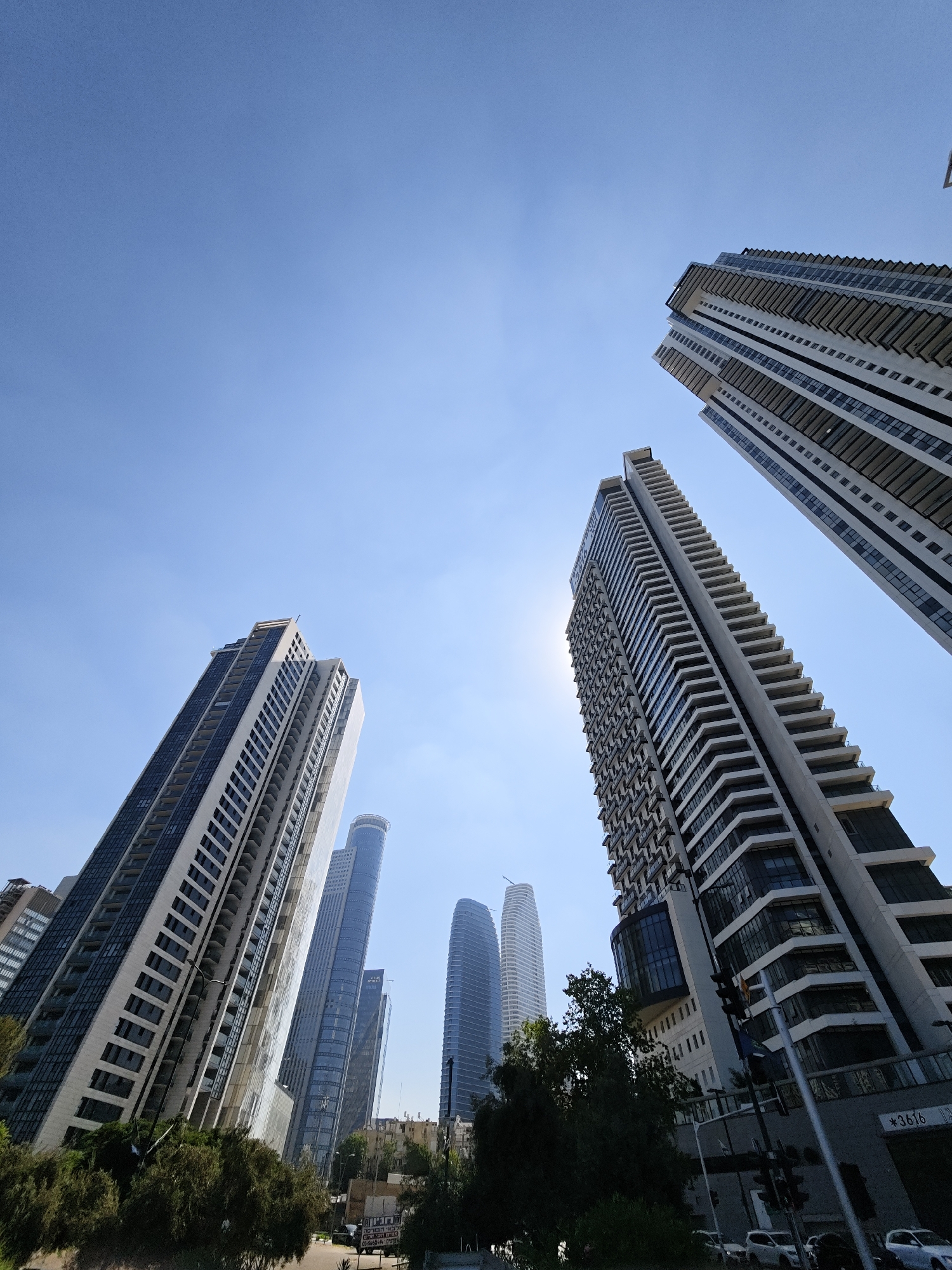 The image shows a low-angle view of several modern skyscrapers against a clear blue sky. The buildings are predominantly white and glass, with balconies and layered architectural features. Some of the skyscrapers appear to be residential towers, indicated by the numerous balconies. In the foreground, there are trees and some street-level infrastructure, including traffic lights and what appear to be cars parked or driving on a street. The overall scene suggests an urban environment with a focus on contemporary architecture. This scene is located in the city of Givatayim, Israel.