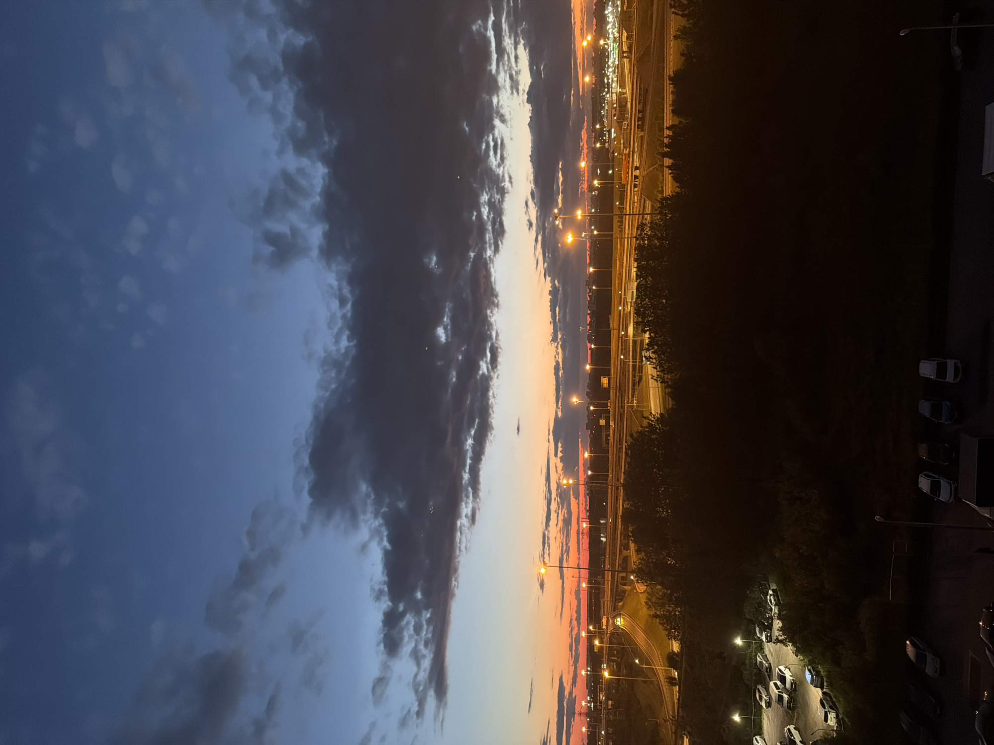 A wide-angle, vertical shot captures a city skyline at dusk, with a dramatic sky gradient transitioning from deep blue at the top to a fiery orange near the horizon. Dark, textured clouds drift across the upper portion of the sky. Below the sky, streetlights illuminate a complex network of highway overpasses and roads, casting a warm glow on the scene. A dense line of trees separates the highway from a dark foreground where several cars are parked in two separate lots. The overall impression is one of urban development meeting the natural transition from day to night.