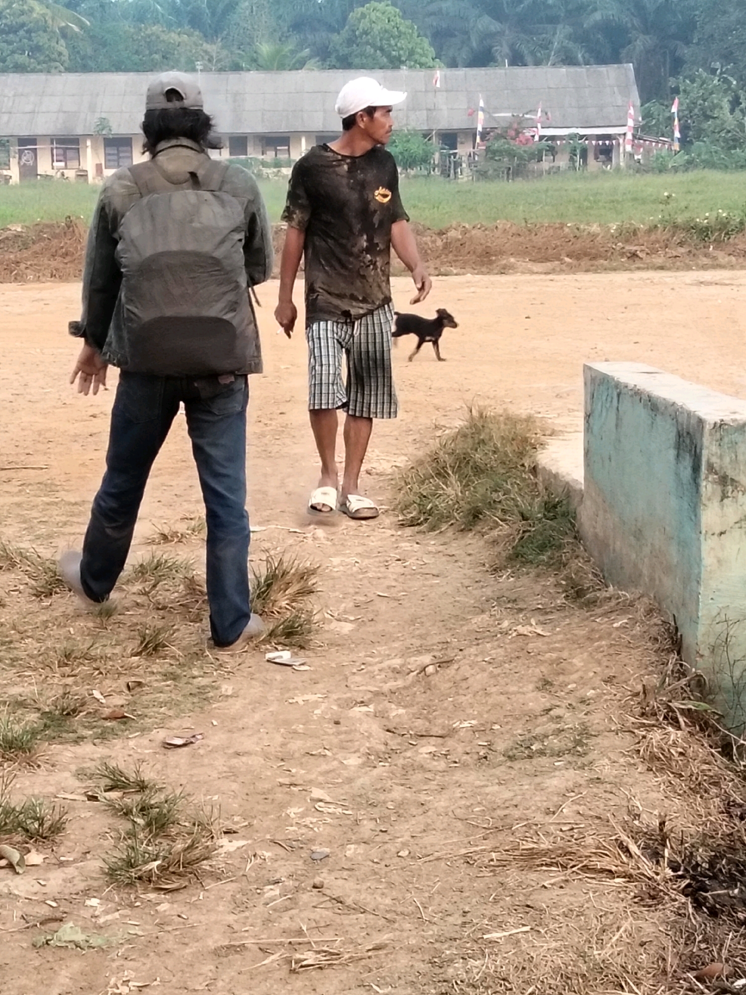 In Teluk Tenggirik, Indonesia, two individuals are observed in an outdoor, unpaved area. The person in the foreground, facing away from the camera, is wearing a grey backpack and blue jeans. To their right, a second person, facing left, is wearing a black t-shirt and plaid shorts. A small black dog is visible between the two individuals, in motion. In the background, a building with a corrugated roof and flags is situated amidst vegetation. A low concrete structure runs along the right side of the foreground. The ground is composed of dry soil and sparse grass.
