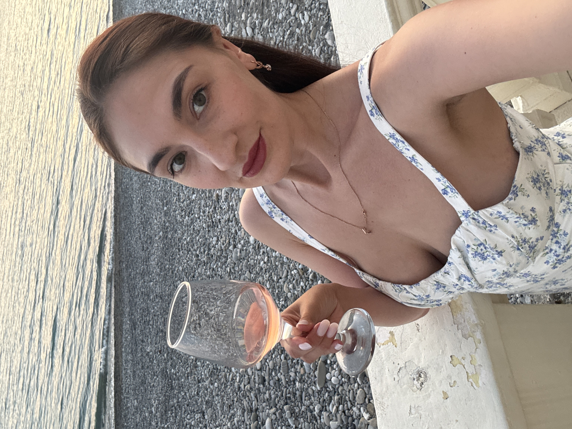 A woman with reddish-brown hair, green eyes, and red lipstick is depicted in a selfie, looking directly at the camera. She has visible freckles on her nose and cheeks. She is wearing a white sundress adorned with blue floral patterns and a delicate gold chain necklace, along with a small gold hoop earring in her right ear. Her right hand, featuring light pink and white French tip nails, holds a stemmed glass containing rosé wine. She is positioned next to a white concrete or stone railing, which shows signs of wear and peeling paint. The background consists of a pebble beach meeting the calm, rippled water of the Black Sea, illuminated by the soft light of evening. The location is the coastal city of Sochi, Russia.