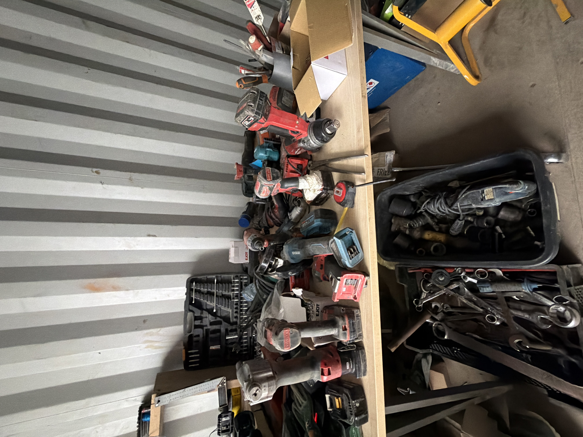 An indoor scene, likely a workshop or storage area, with the ribbed metal walls of a shipping container forming the background. A wooden shelf extends across the middle of the image, cluttered with various power tools, toolboxes, and other equipment. Prominently displayed are several cordless drills and impact wrenches, many with red and black casings, along with their associated batteries. To the left, a black toolbox is open, revealing a set of wrenches and sockets. Below the shelf, a dark bin contains more tools, including wrenches and what appears to be a small rotary tool. A yellow ladder-like object is visible on the right side of the image. The overall impression is one of organized chaos, suggesting a working environment for repairs or construction. The location is identified as Janowice, Poland.