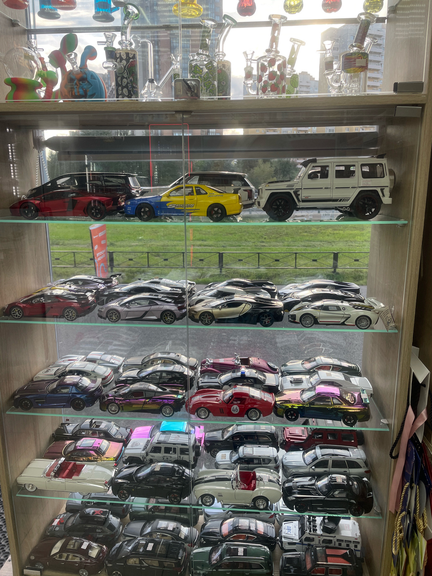 The image displays a glass-enclosed display case filled with a collection of model cars and decorative glass items. The display case is situated in what appears to be a retail establishment, possibly a gift shop or a specialty store. The top shelf showcases a variety of colorful glass pieces, some with intricate designs and shapes, including what appear to be smoking accessories. Below this, several shelves are meticulously arranged with a diverse assortment of model cars. These models range in style and color, featuring sports cars, SUVs, and classic vehicles, all presented in a static, organized manner. The background visible through the display case suggests an outdoor environment with greenery and a fence, indicating the store is likely located in an urban or suburban area. The lighting suggests it is daytime, with sunlight potentially illuminating the scene from the left. The overall impression is one of a curated collection, presented for viewing and potential purchase. There are no people visible in the image. Without additional context or external information, the specific location in Saint Petersburg, Russia, cannot be determined from the image alone.