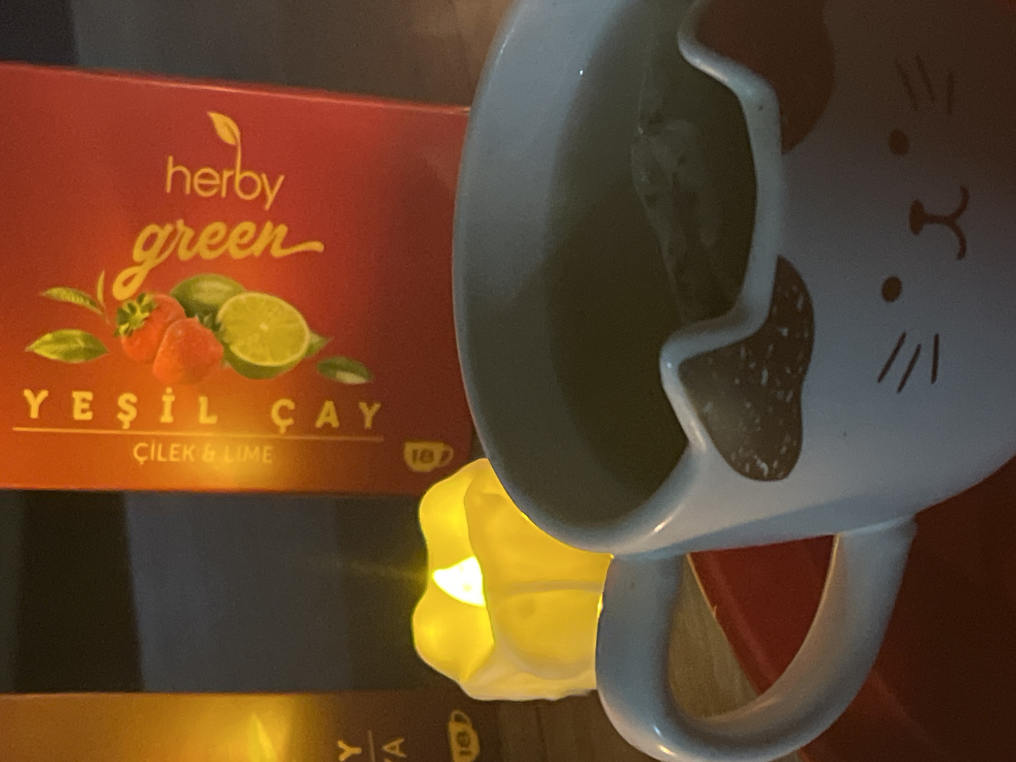 The image shows a box of Herby Green "Yeşil Çay" (Green Tea) with strawberry and lime flavor, next to a lit, artificial candle, and in front of a mug with a cat face design. The text on the tea box is written in both English and Turkish. The image was taken indoors with soft, warm lighting. There's no indication of Alanya, Turkey in the image.