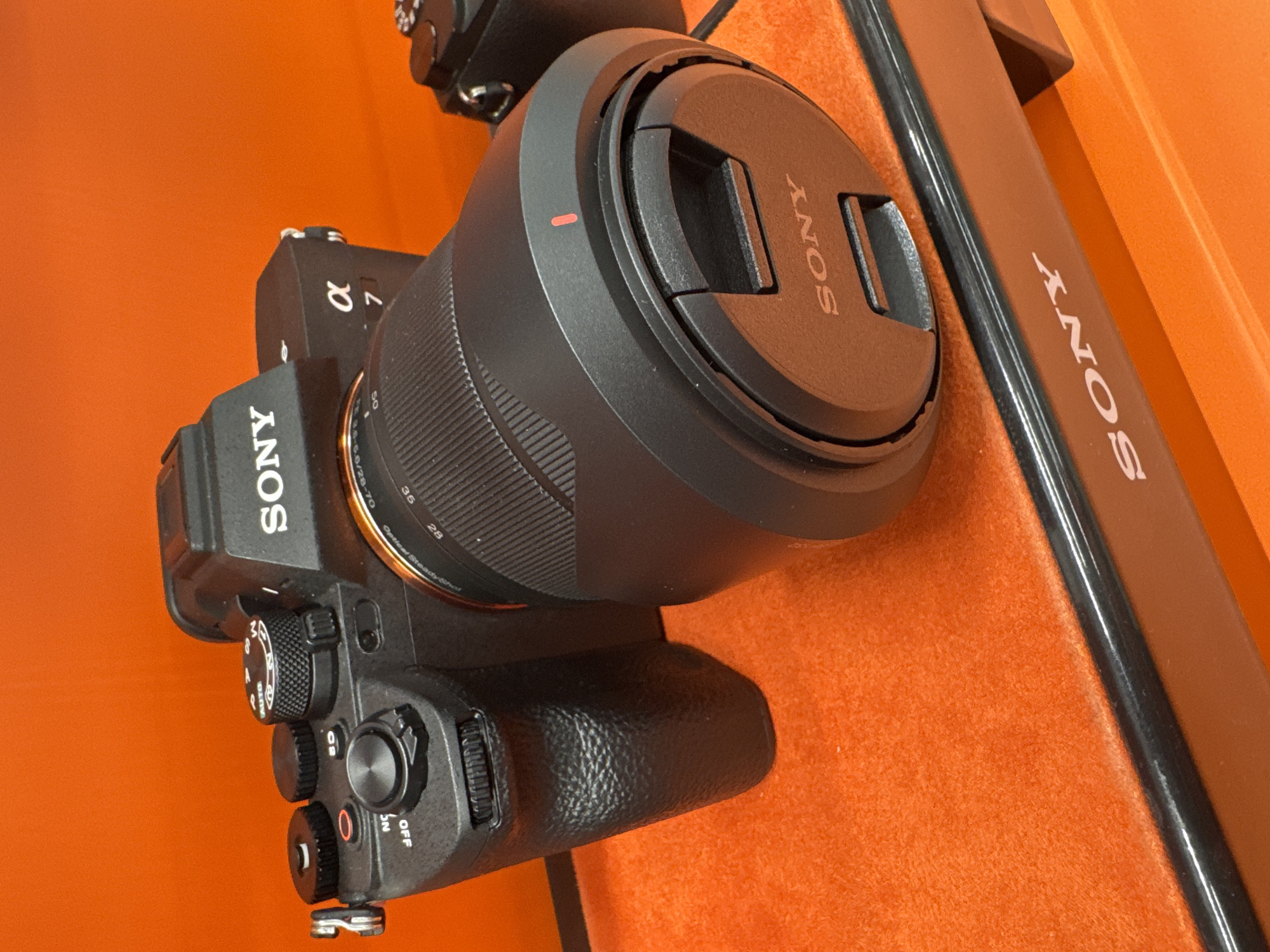 The image shows a black Sony α7 camera with a lens attached, and the lens has a lens cap on. The camera is placed on an orange surface next to a long, black Sony display plaque with the Sony logo printed in white text. The camera's dials and buttons are visible, including the power switch in the "OFF" position. The camera lens indicates a focal length range of 28-70mm and aperture settings. The setting is indoors, and based on the product placement, it could potentially be in a retail environment that carries Sony products. There is no specific indication in the image that suggests that the location is Sofia, Bulgaria.