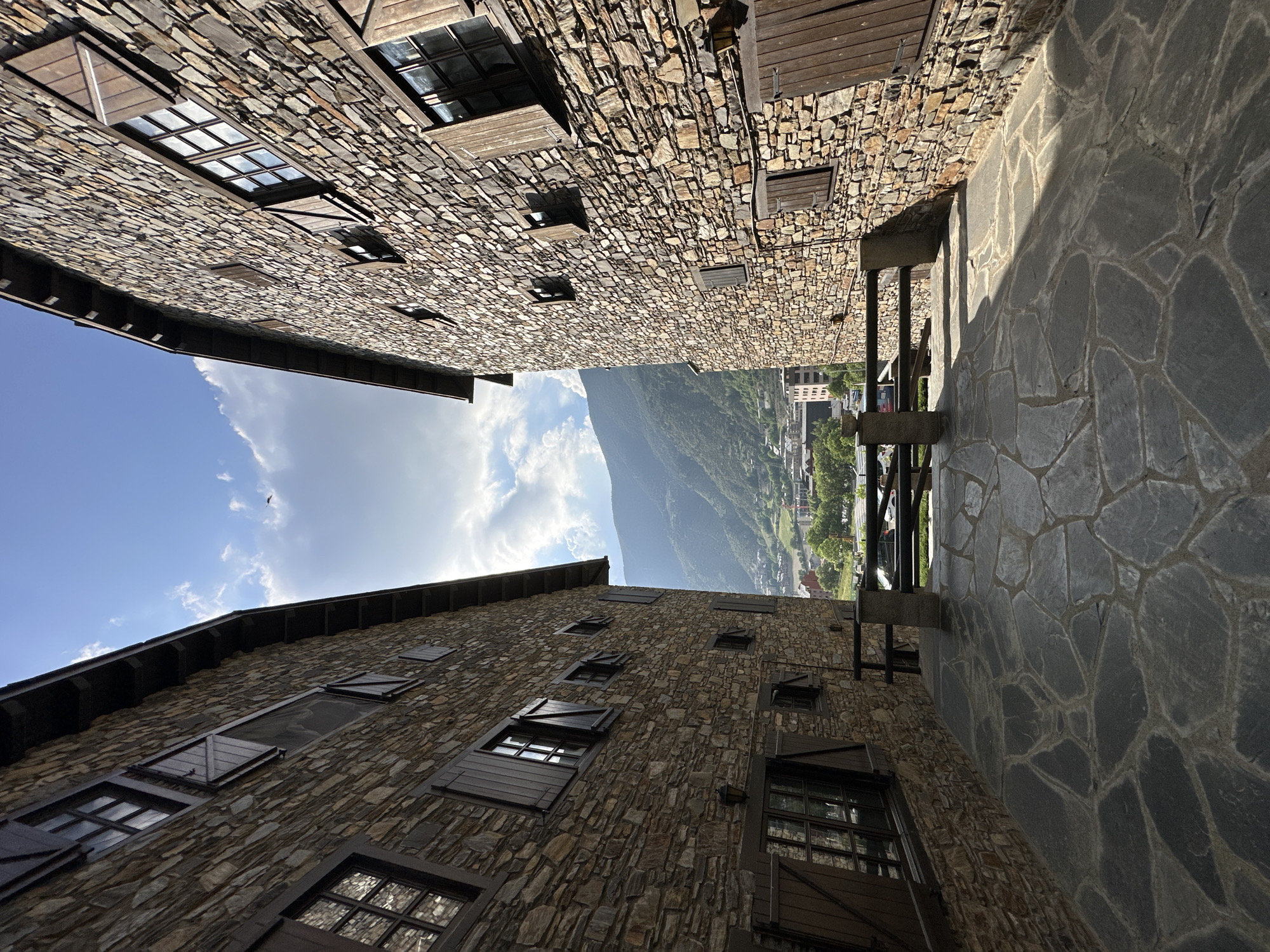 The image displays a narrow passage or alleyway in Encamp, Andorra, framed by two traditional stone buildings on either side. Both structures feature facades built from irregular, multi-toned stone blocks and multiple windows with dark wooden frames and shutters, some of which are open.
The ground of the passage is paved with large, irregularly shaped flagstones, creating a textured surface. Strong sunlight illuminates parts of the scene, casting distinct shadows on the paving and the building to the right, suggesting it is daytime, likely late morning or early afternoon. A dark wooden railing or fence-like structure is visible along the side of the right-hand building.
Beyond the confined space of the alley, the scene opens up to a vast mountainous landscape. A prominent, green mountain dominates the background under a bright blue sky dotted with white clouds. In the valley below the mountain, glimpses of a town, including various buildings and green trees, can be seen, characteristic of the region.
The perspective is upward-looking, from a low vantage point within the alley, emphasizing the height of the buildings and the scale of the distant mountain. There are no people visible, and no visible text in the image.