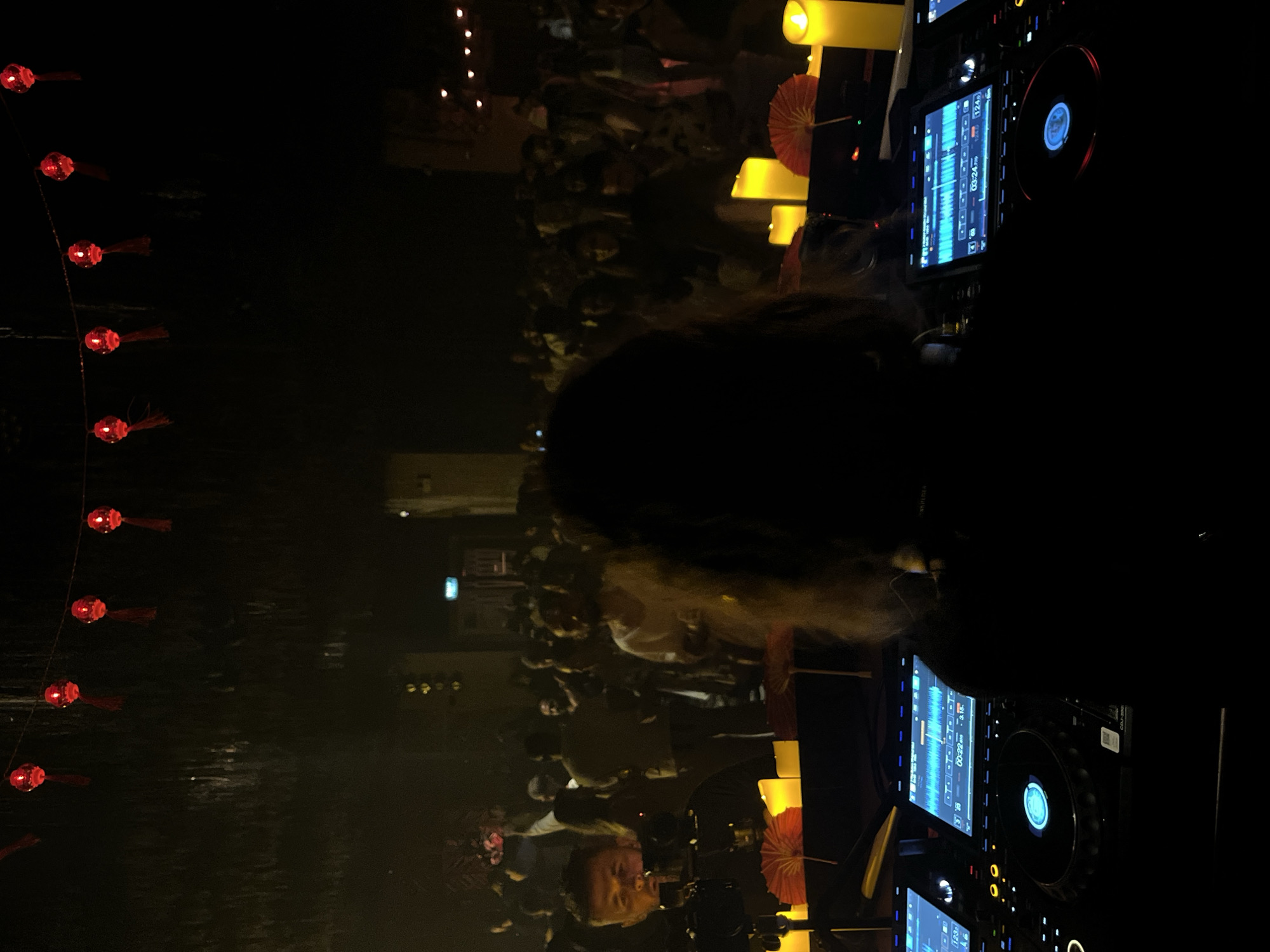 This is a dimly lit, vertical photograph taken at night, possibly outdoors or in a venue with an open-air feel, given the presence of what appears to be water in the foreground. The scene is dominated by a crowd of people, mostly silhouetted against a dark background. In the left foreground, a string of red lanterns hangs, their light illuminating the dark water below, creating ripples.
Central to the image, a DJ is visible, their head and shoulders in silhouette, facing away from the camera. They are positioned behind DJ equipment, including turntables and digital screens displaying waveforms and track information. One screen shows "03:24". To the right of the DJ, several large, bright yellow cylindrical lights, possibly candles or illuminated decorations, are prominent, casting a warm glow. Red paper umbrellas or decorative fans are also visible near the lights.
The crowd in the background appears to be enjoying themselves, with some individuals leaning in and gesturing. The overall atmosphere suggests a lively event, possibly a music performance or party. The time of day is clearly night, and while no specific weather is indicated, the presence of water suggests a potentially humid environment. The red lanterns and paper umbrellas hint at an Asian cultural influence, fitting with the provided location context of Ban Bang Thao, Thailand. The emotions conveyed are generally positive and energetic, driven by the music and the gathering.