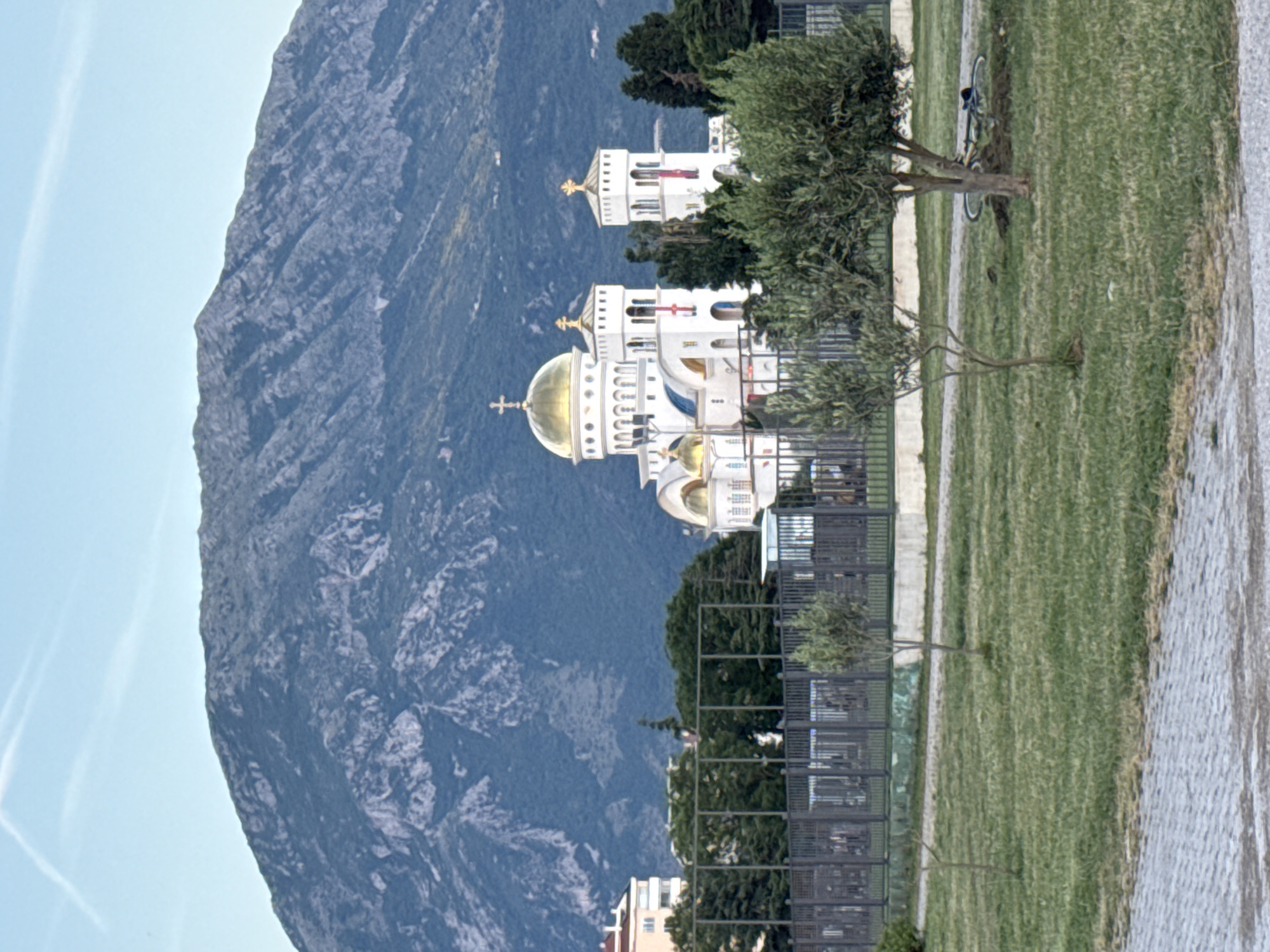 This image depicts an urban landscape in Bar, Montenegro, dominated by religious architecture and a natural mountainous backdrop. In the background, a large, dark grey-blue mountain range with visible rocky outcroppings and sparse vegetation extends across the horizon under a clear, light blue sky. A single, faint white contrail is visible in the upper left quadrant of the sky.
The midground is characterized by the prominent St. Jovan Vladimir Orthodox Cathedral, a large white structure featuring multiple golden domes topped with crosses. The central dome is the largest, flanked by smaller golden domes and two tall, square white bell towers, each adorned with a red banner hanging vertically. To the left and right of the church, various green trees and other light-colored multi-story buildings are visible. A dark metal fence, possibly enclosing a park or sports complex, stretches horizontally across the midground in front of the church and other buildings.
The foreground consists of a section of a grey paved pathway made of interlocking pavers on the left, transitioning into a somewhat patchy green grass field that covers the majority of the lower image. Two young olive trees with slender trunks are planted in the grass. A bicycle, predominantly dark-colored, lies on its side in the grass next to the base of the larger olive tree on the right. No individuals are present in the scene. The lighting suggests daytime conditions.