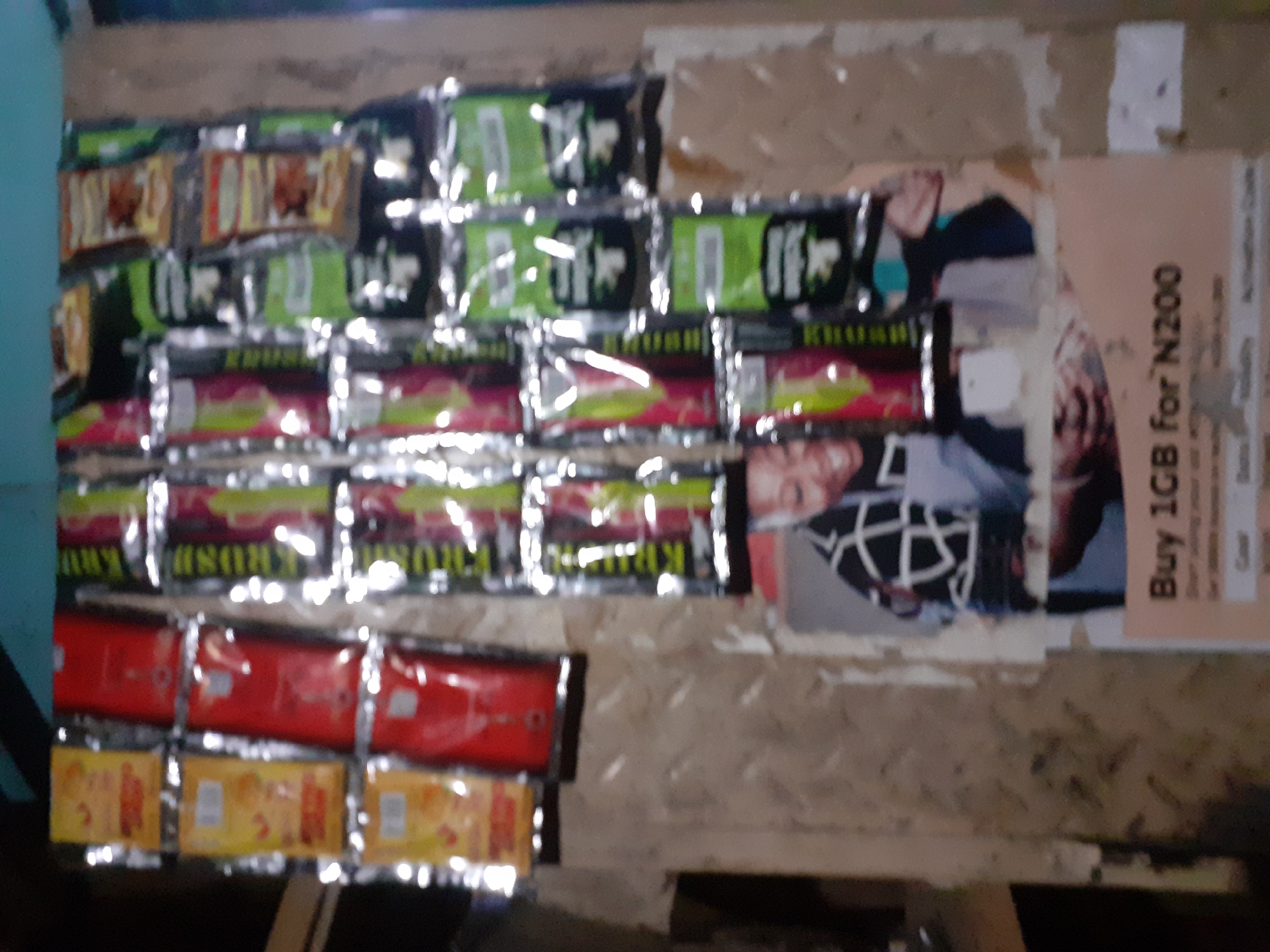 The image depicts a close-up view of a street vendor's stall in Jalingo, Nigeria. The stall is laden with various small, packaged goods, predominantly snacks or confectionery, displayed in rows. The packaging is colorful, featuring shades of yellow, red, and green. In the lower portion of the image, a promotional poster is visible, advertising "Buy 1GB for N200". The poster features a portrait of a smiling individual, likely a model, wearing a black and white checkered garment. The overall environment suggests an outdoor market setting, with the stall constructed from what appears to be wood or corrugated metal. The lighting indicates it is daytime, with strong highlights on the plastic packaging of the products, suggesting a somewhat bright and possibly humid atmosphere.
