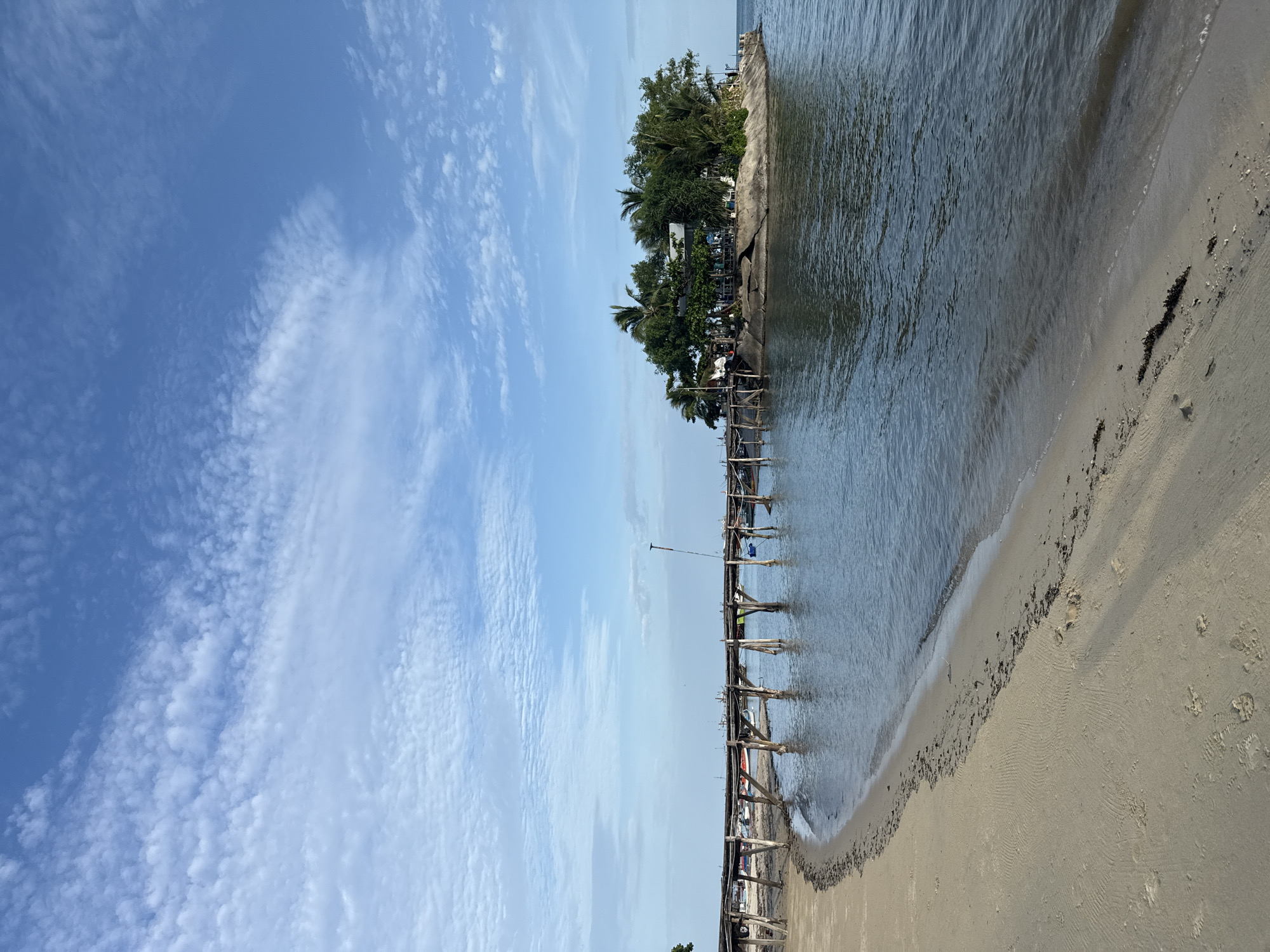 The image depicts a serene coastal scene in Ko Samui District, Thailand. A wooden pier extends from a sandy beach towards a small, vegetated island in the ocean. The island is topped with lush trees and a building, suggesting it might be a resort or a local establishment. The water is calm, reflecting the blue sky dotted with scattered white clouds. Several boats are visible near the island and along the shoreline, indicating local fishing or transportation activities. The beach is marked with footprints and some seaweed, hinting at recent human presence. The overall ambiance is tranquil and picturesque, characteristic of a tropical island setting.