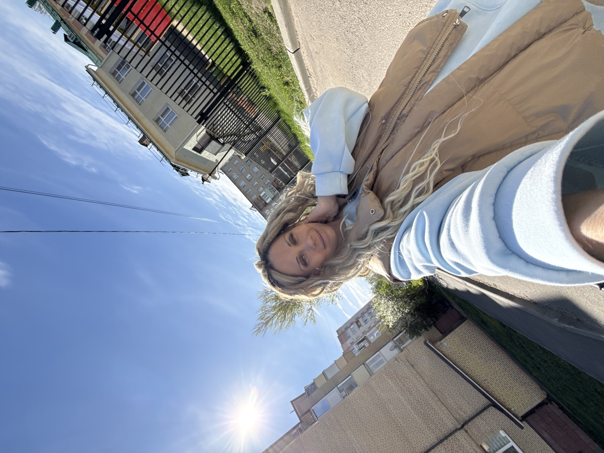 A selfie image shows a woman on a sunny day in Заволжье, Russia. She is wearing a light blue hooded sweatshirt and a tan vest. The background includes apartment buildings, a fence, a sidewalk, and a bright blue sky with the sun shining. The image appears to be taken at an angle.