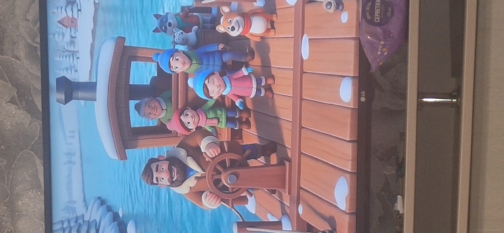 The image depicts a digitally rendered scene of a group of cartoon characters on a wooden dock next to a body of water, likely a river or lake, during winter. The characters include a man with a beard at a ship's wheel, an elderly woman in the background of a small wooden cabin, two children, and two animals, possibly a fox and a dog. They are all dressed in winter clothing. The environment is snowy, with trees and a building visible on the distant shore. A purple bag with Russian text is visible in the lower right corner of the image, resting on a shelf below the screen displaying the scene. There is no information in the image or the provided context that indicates the location is in Bila Tserkva, Ukraine.