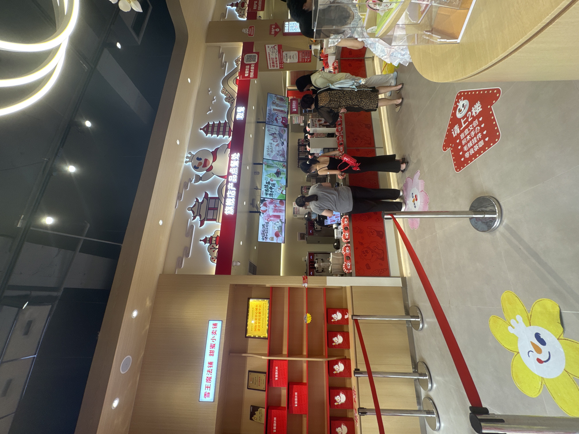 Here is a detailed description of the image:
**Scene:** The image shows the interior of a Mixue (蜜雪冰城) store. Mixue is a popular Chinese tea and ice cream chain. There is a line of people waiting to order.
**Location:** Based on the writing and signage, the location is likely in the Nan'an District (南岸区) of China, possibly in a city with a designated international consumer center.
**People:** Several people are standing in line at the counter. They appear to be customers waiting to place their orders. Their clothing is casual. A person behind the counter appears to be an employee.
**Objects:**
* **Menu Screens:** There are digital menu screens displayed above the counter showing various drink and dessert options offered by Mixue.
* **Shelving:** There is shelving along the wall displaying various merchandise, including red boxes with the Mixue snowman logo.
* **Queue Barriers:** Queue barriers with red straps are used to organize the line of customers.
* **Signage:** There is various signage with Chinese characters, including promotional material and instructions for ordering. One sign indicates a second floor with "Blind Box Cultural & Creative" and "Snack Tea and Coffee."
* **Floor Decals:** There are decals on the floor, one of which is a drawing of a flower, and another is an arrow pointing upstairs to a second floor.
**Actions:** Customers are waiting in line to place their orders. The employee behind the counter is presumably taking orders and preparing drinks.
**Interactions:** The customers are waiting in line. They are likely reading the menu to decide what they want to order.
**Environment:** The store appears to be clean and well-lit. The color scheme is mainly red and white. The store's design incorporates cartoon-like elements, including the Mixue snowman logo.