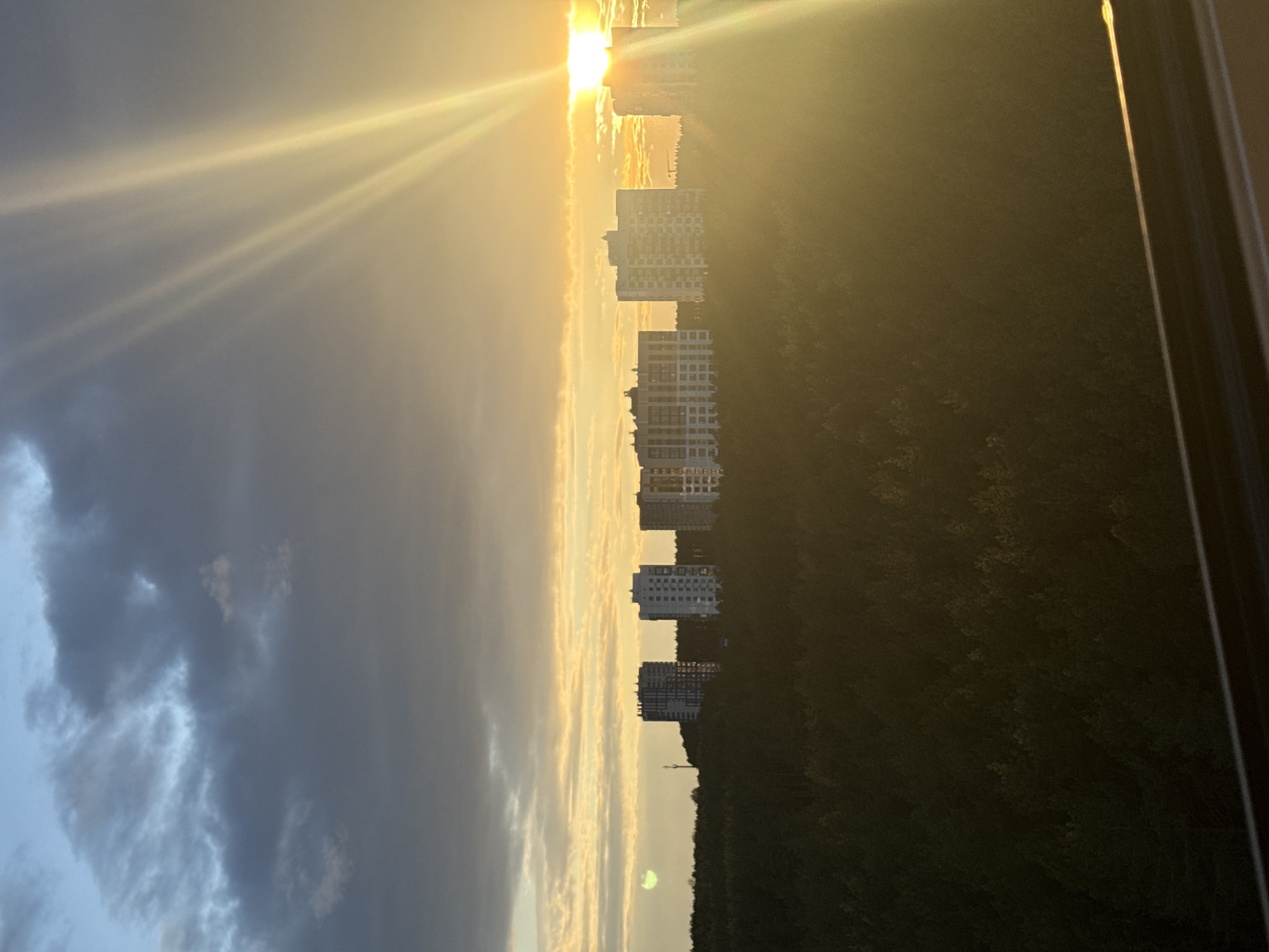 A dramatic landscape photograph captures a sunrise or sunset over a suburban or urban edge. The sun, a brilliant golden orb, is positioned on the right side of the frame, low on the horizon, radiating prominent, long rays that stretch diagonally across the sky.
The sky exhibits a clear gradient: dark, turbulent clouds occupy the upper left portion, transitioning into warm golden and orange hues closer to the sun. Below the illuminated sky, a horizontal line of several modern high-rise residential buildings stands. These white or light-colored buildings are partially silhouetted or subtly lit by the low sun, with visible windows and balconies. Beneath the buildings, a dense, dark green forest or wooded area forms a natural boundary, stretching across the lower part of the scene.
The image appears to have been taken from an elevated indoor viewpoint, as a dark window frame or sill is visible along the far right edge, hinting at the perspective. The overall atmosphere is serene yet dramatic, emphasized by the strong contrast between the bright sun and the shadowed foreground and darker clouds. There are no people or discernible text visible in the image.