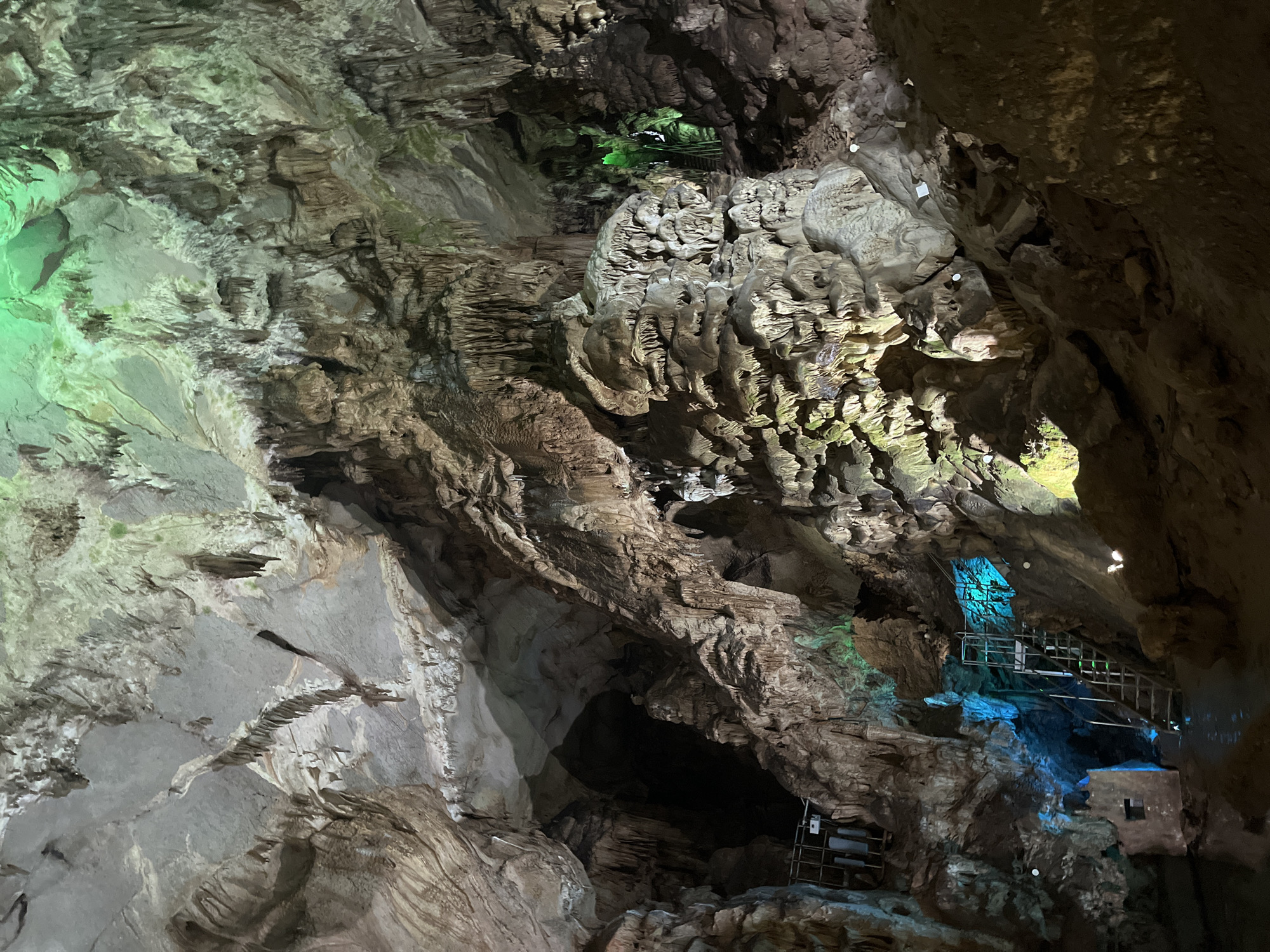 The image shows the interior of a cave, likely a tourist attraction, with various speleothems illuminated by artificial lights. The cave ceiling and walls are covered with stalactites and stalagmites of various shapes and sizes, some appearing as delicate icicle-like formations, while others are massive and columnar. The rock formations are predominantly earthy brown and grey, with some areas tinged green from moss or algae, and others highlighted by colored lights.
In the lower left portion of the image, a metal staircase with handrails is set up, ascending into the cave. The steps are lit by a bright blue light source, illuminating the water pool at the base of the stairs and the surrounding rock faces. Further into the cave, a section of the rock formation is bathed in green light, highlighting its textured surface. In the background, the cave opens up into darker recesses, suggesting a larger and more extensive underground space. The overall impression is one of natural geological formations enhanced by lighting for visual effect, likely within a subterranean tourist destination.