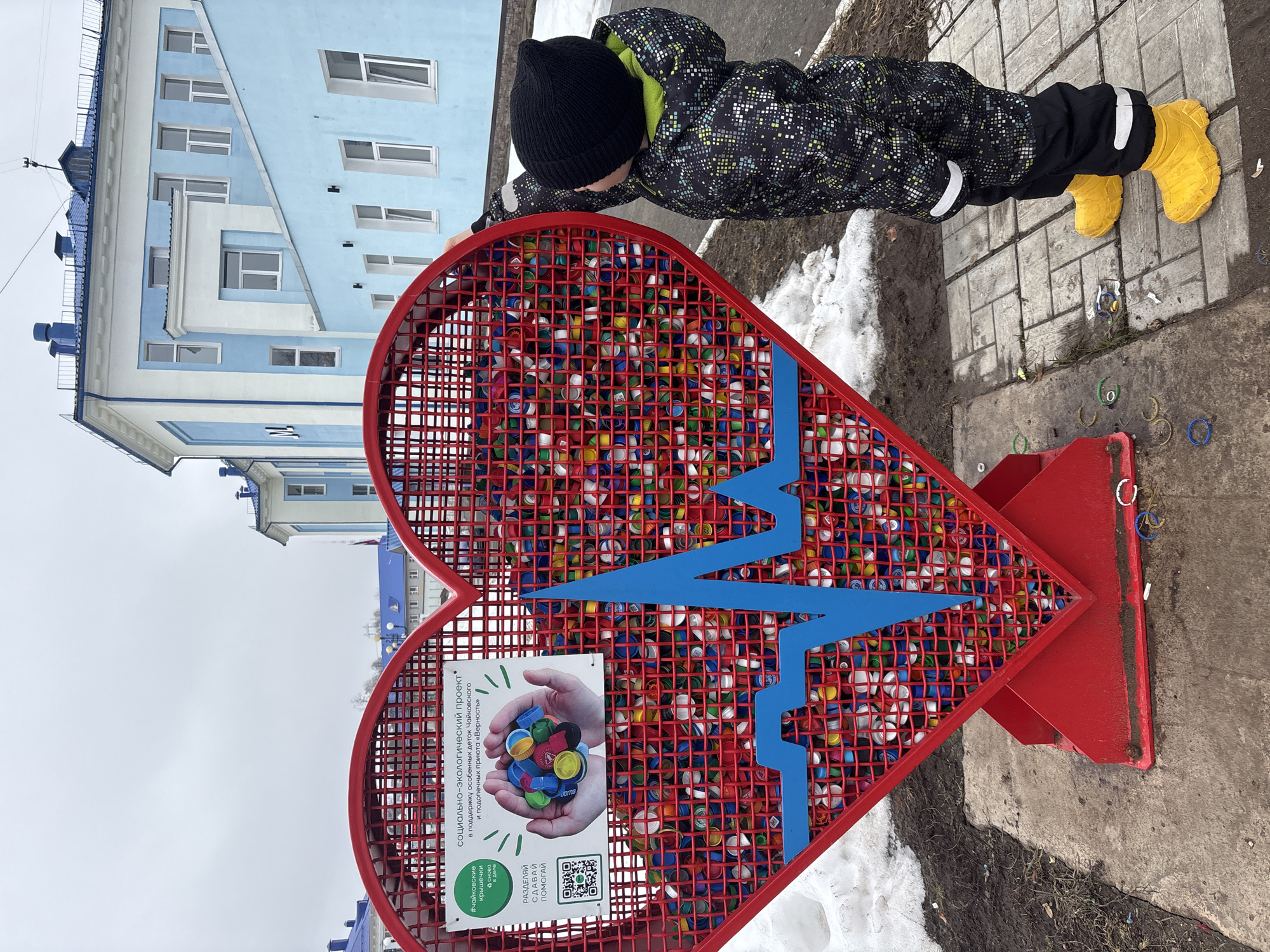 A child in a black hat, patterned winter coat, black pants, and yellow boots leans over a large, red, heart-shaped public art installation made of metal grid and filled with colorful plastic bottle caps. A blue lightning-bolt design is painted across the surface. A sign attached to the heart explains the project as a social-ecological initiative in support of special needs children in Chaikovsky, Russia, and clients of the "Verznost" shelter. Snow patches remain on the ground. In the background, there is a multi-story, light-blue building. The surrounding ground is concrete.