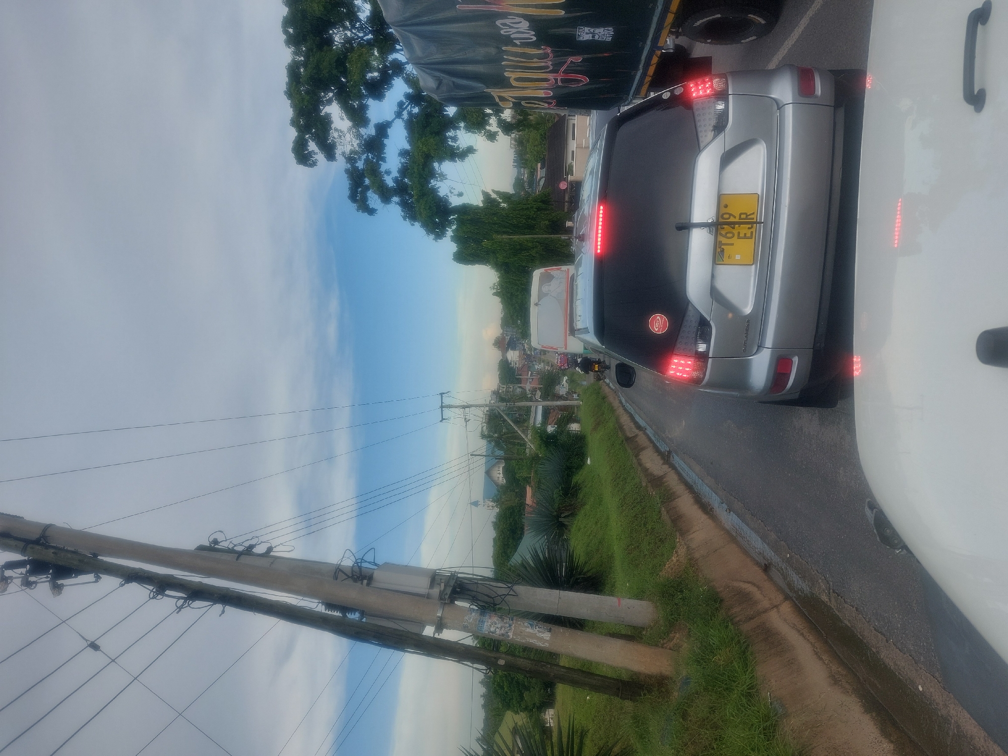 The image depicts a traffic scene in Dar es-Salaam, Tanzania. Foreground features the back of a silver car with license plate "T629 EJR," followed by a white vehicle. In the background, a truck with illegible writing is visible. In the mid-ground, the landscape includes greenery, houses, and a church steeple. A utility pole with wires is on the left side of the frame. The sky is a mixture of blue and cloudy conditions.