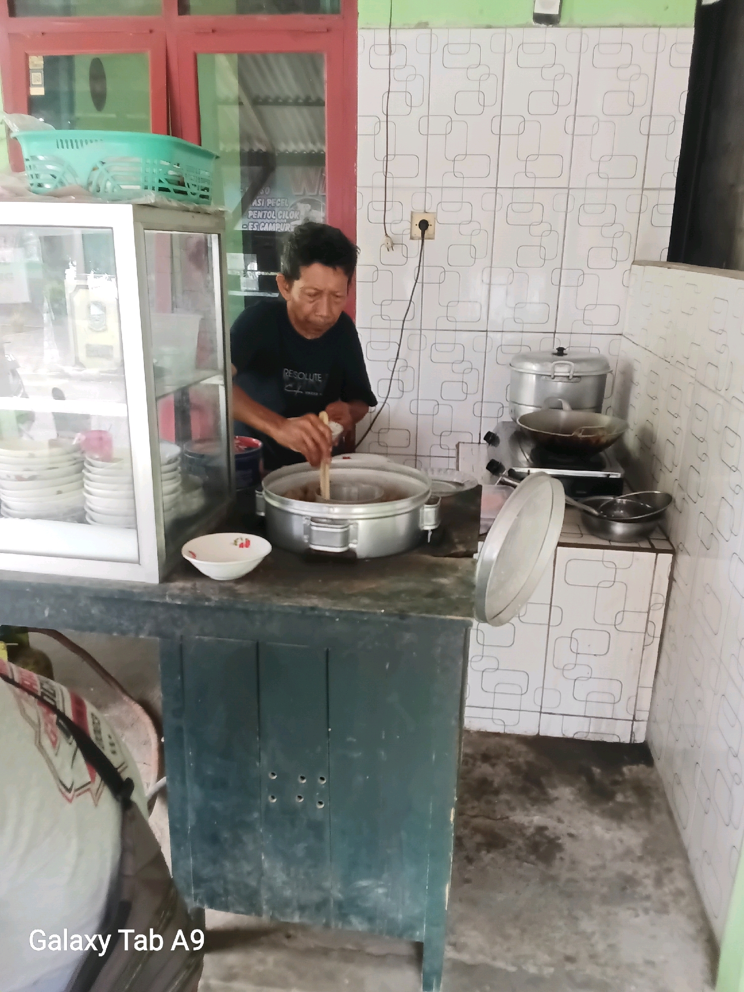 A man in a black t-shirt is stirring food in a large silver pot on a stove. He is standing in a kitchen with white tiled walls that have a repeating geometric pattern. To his left, a glass display case contains stacks of white plates. In front of the display case, a small white bowl with a floral design sits on a dark green counter. Behind the man, a large silver steamer pot is stacked on top of a wok on a gas stove. The floor is concrete and appears slightly wet in places. The scene is likely a small eatery or food stall.