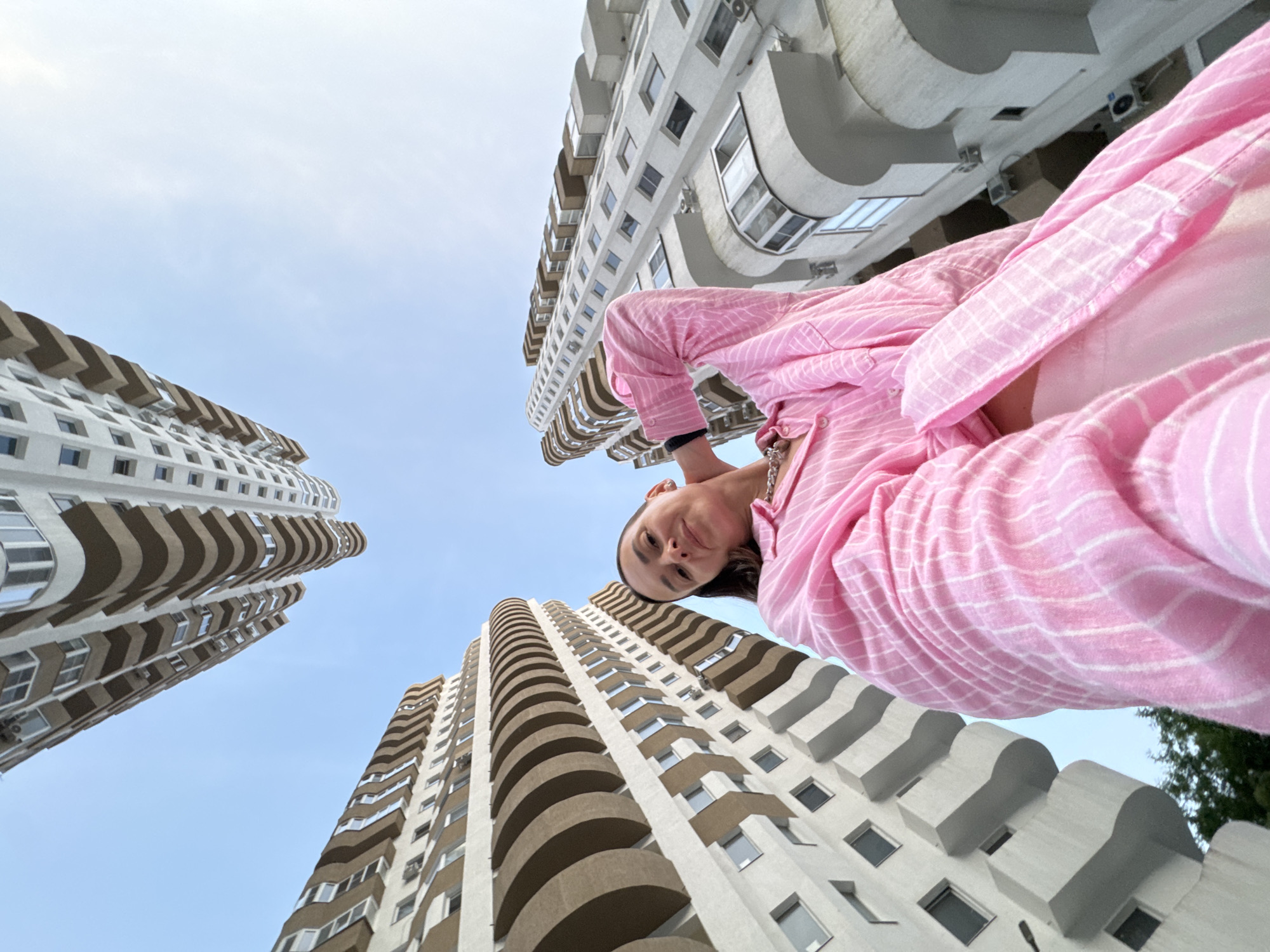 An upward-angle medium shot captures a woman in a pink striped shirt standing against a backdrop of tall, modern apartment buildings in Sochi, Russia. The architecture features curved balconies and a mix of white and brown facades. The sky is a clear, light blue. The woman is looking towards the camera, with her right hand touching her neck, and her left arm is bent. The perspective emphasizes the height of the buildings, making them appear to converge at the top of the frame.